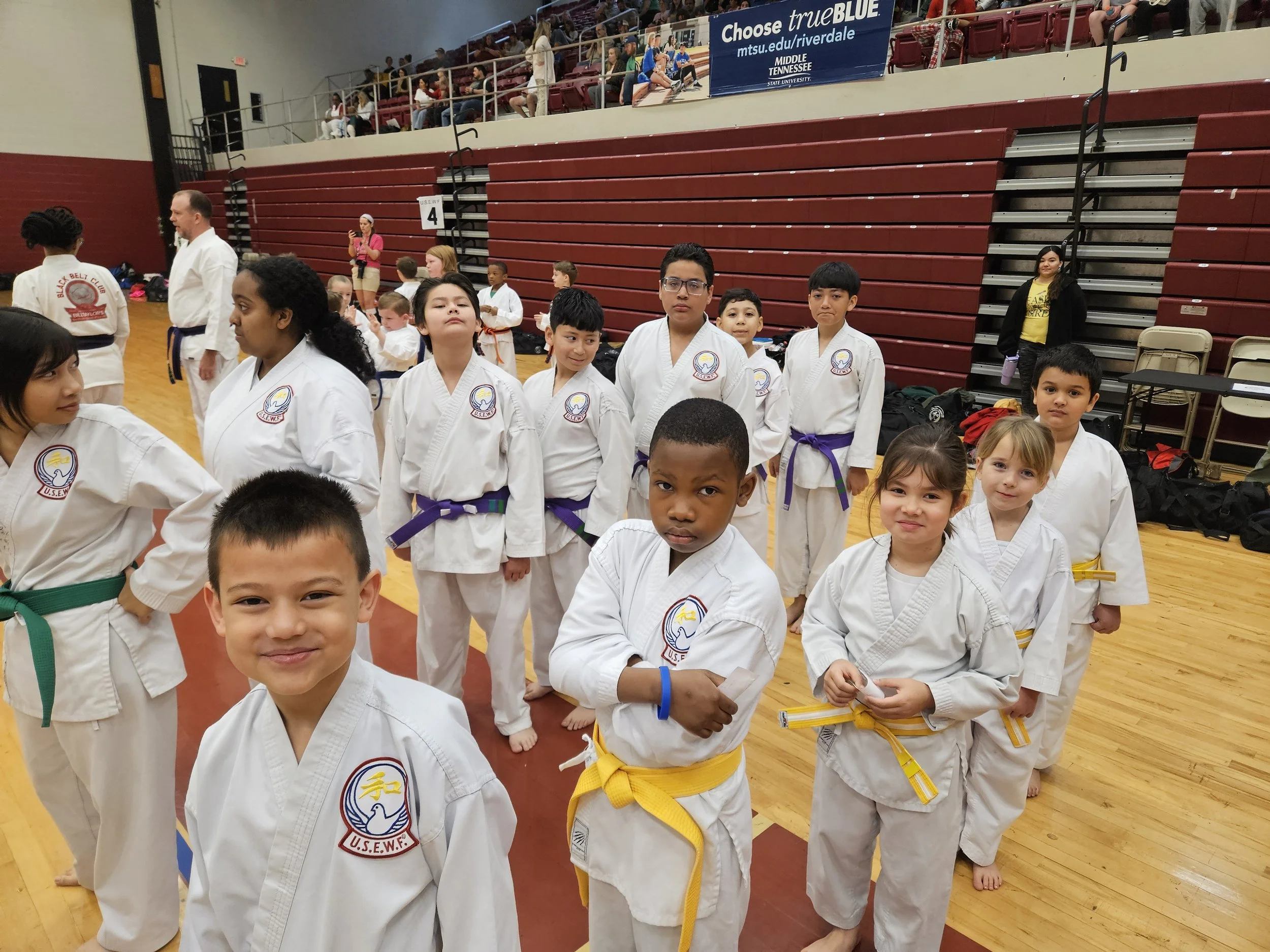 Young children in karate uniforms with colored belts standing in a gymnasium, some smiling and some serious, with spectators in the background and a banner reading 'Choose true BLUE' hanging above.