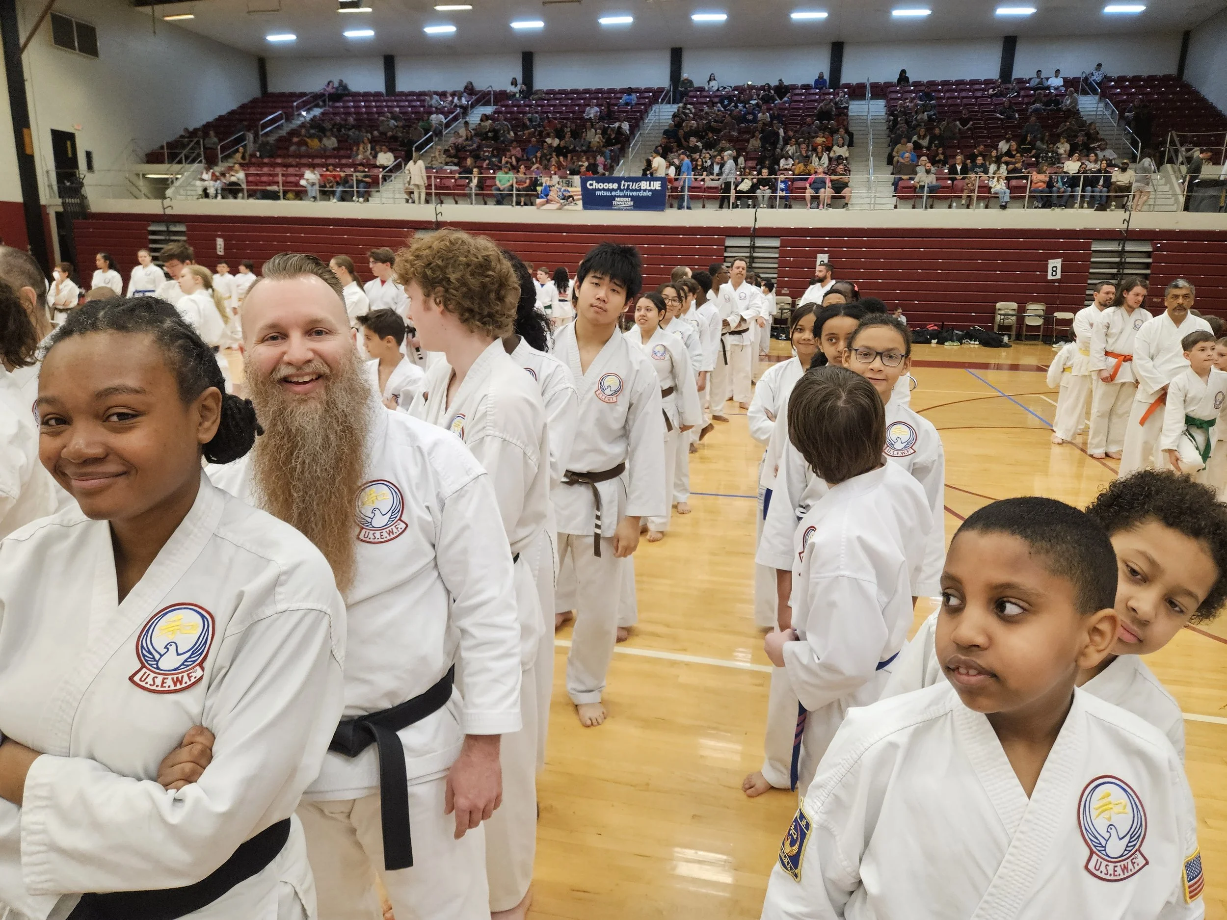 Children and adults in martial arts uniforms lined up inside a gymnasium for a class or event, with a crowd sitting in the bleachers above.