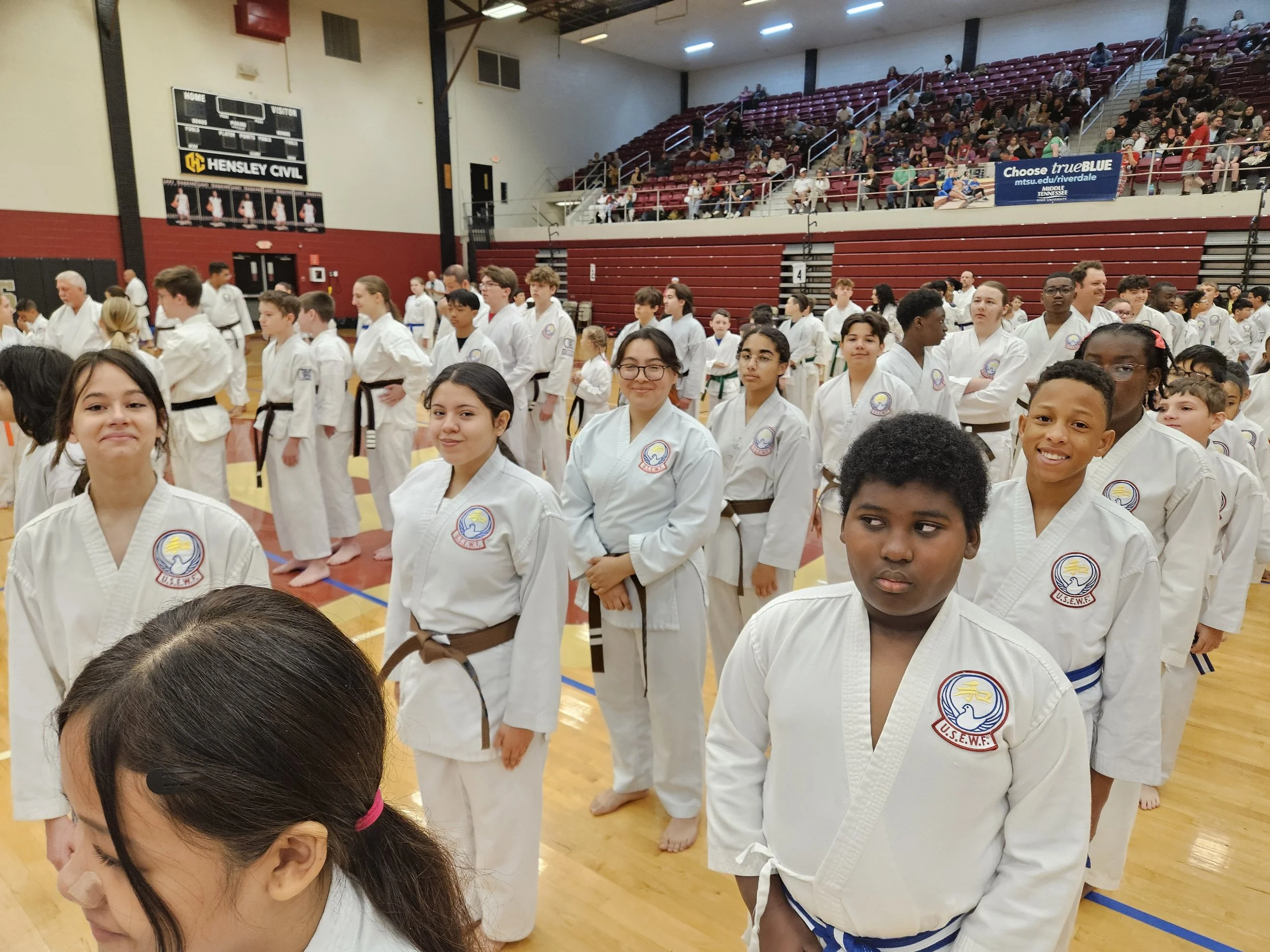 Children and teenagers in martial arts uniforms standing in line inside a gymnasium during a competition or event.