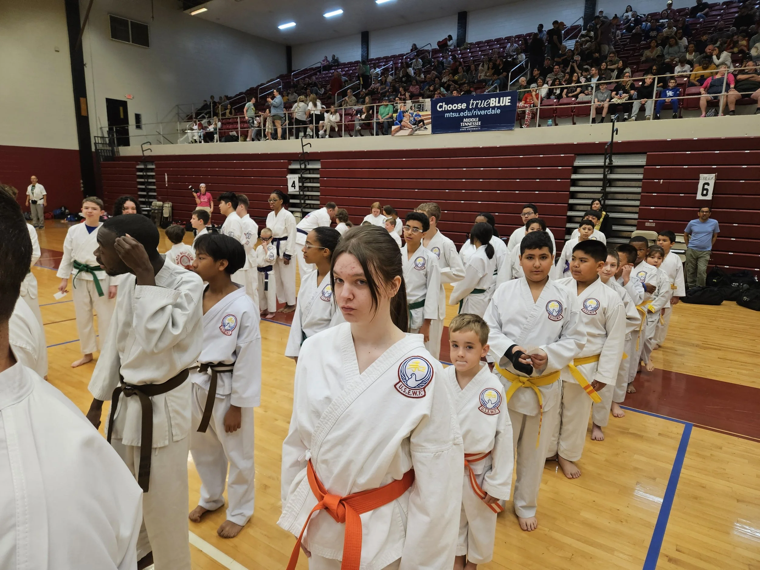 A group of children and teenagers wearing martial arts gis and colored belts, standing in line on a gymnasium floor during a martial arts event with spectators watching from the bleachers.