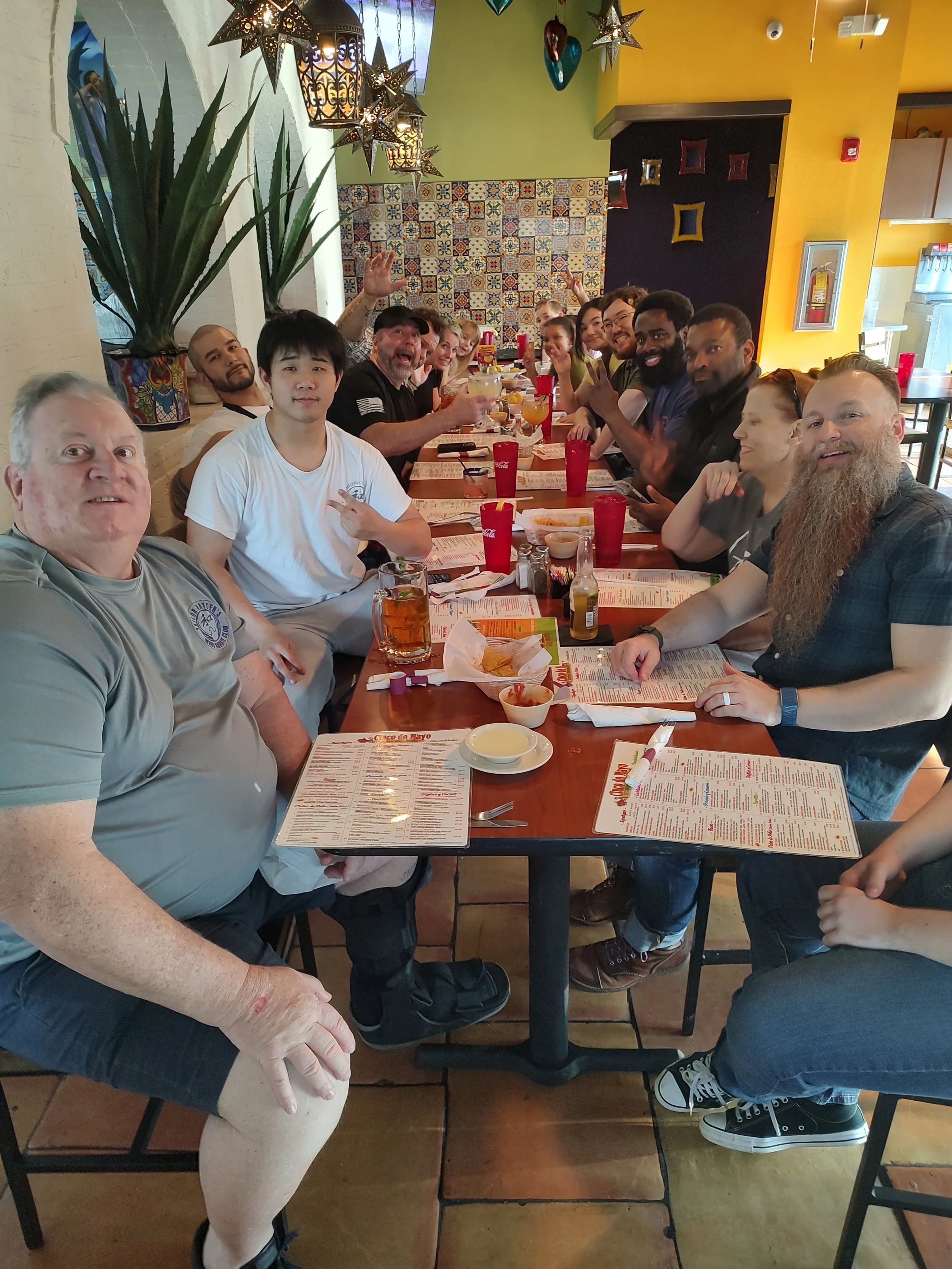 A group of people sitting at a long restaurant table, smiling and raising glasses for a toast. The table has drinks, appetizers, and menus. The restaurant has colorful decor with hanging star-shaped lights, tiled wall, yellow and black painted walls, and large potted plants.