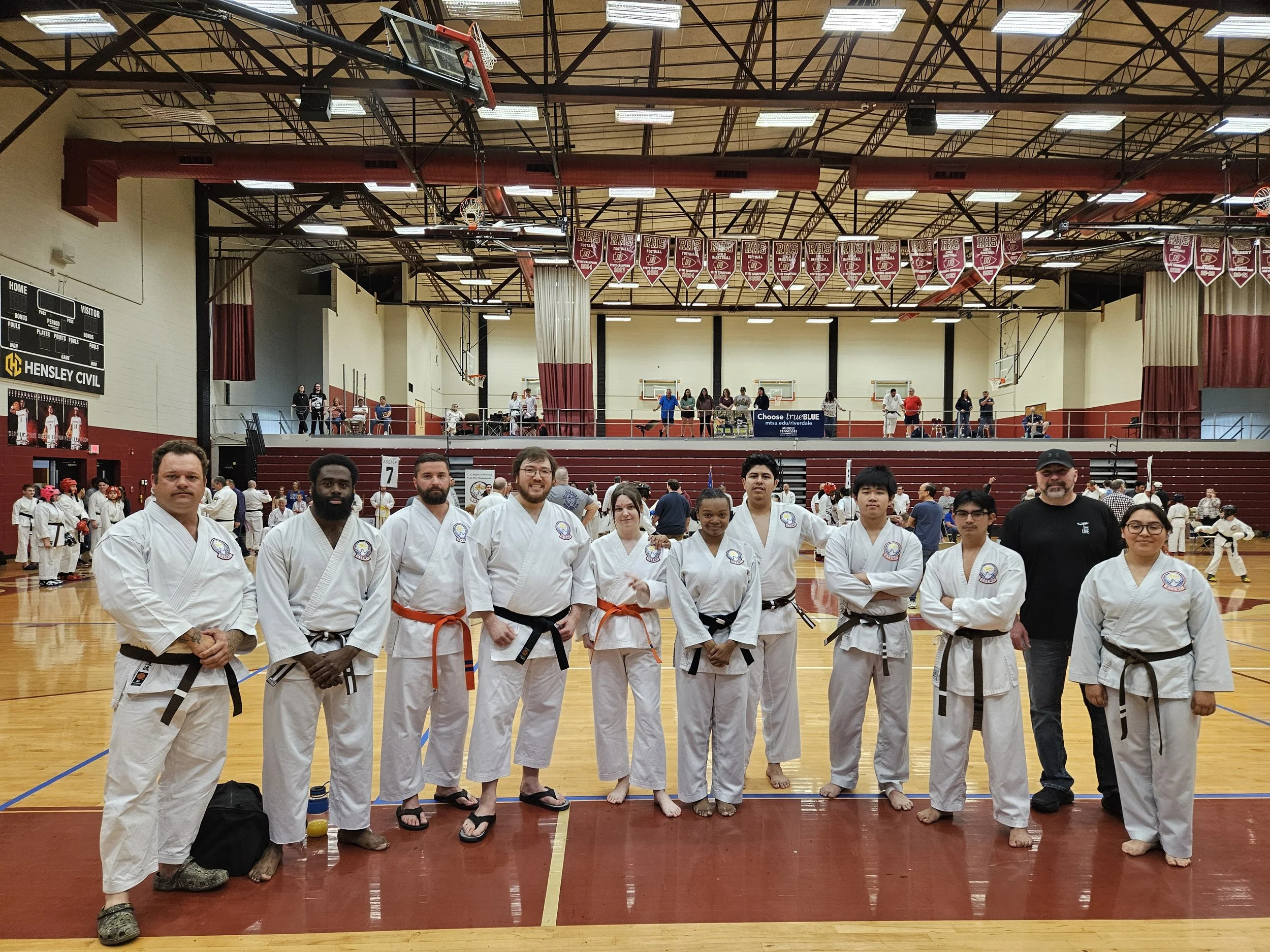 A group of martial artists in white gis with different colored belts, standing in a gymnasium.
