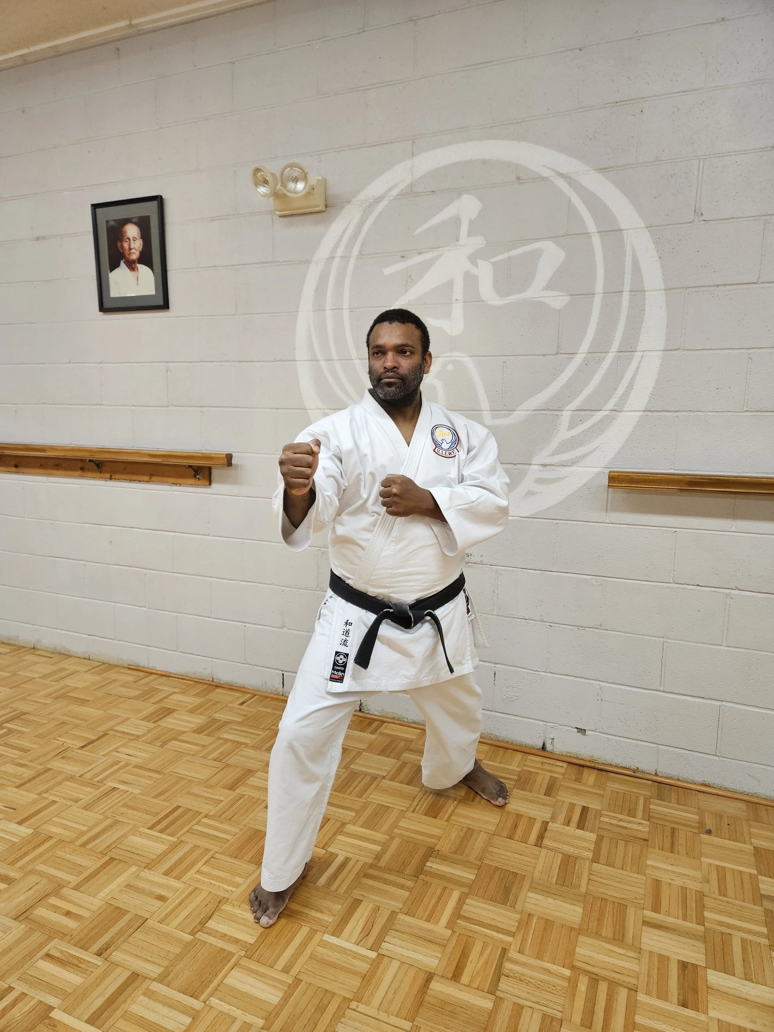 A man wearing a white judo gi with a black belt poses in a martial arts dojo with his fists raised in a fighting stance. The dojo has a wooden floor, a white brick wall, a portrait on the wall, and Japanese calligraphy in the background.