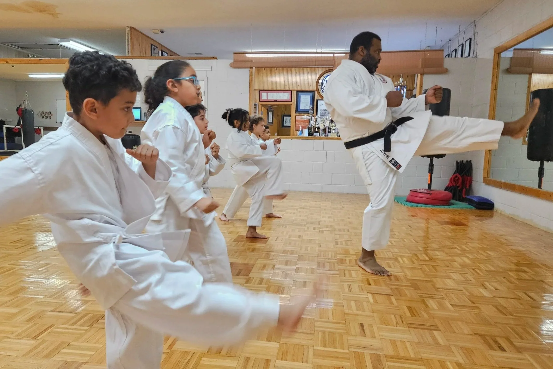 A group of children and an instructor practicing martial arts in a dojo. The instructor is performing a high kick, while the children are doing different martial arts stances, all dressed in white gis.
