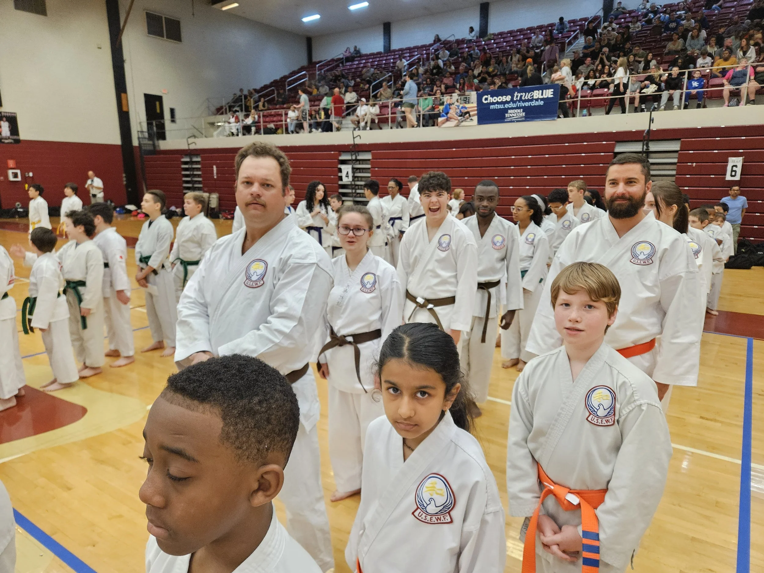 Group of karate students and instructors standing in a gymnasium, dressed in white uniforms with belts, participating in a martial arts event.