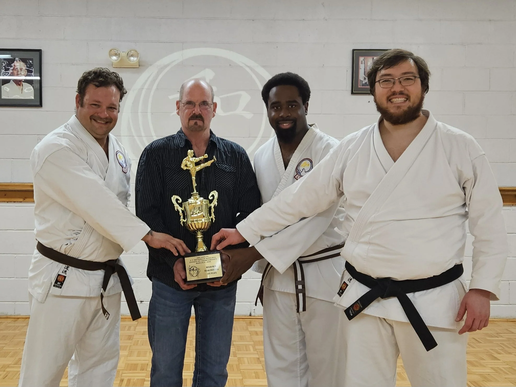 Four martial artists in white uniforms with black belts holding a trophy, standing with a man in casual clothes, inside a dojo with framed pictures on the wall.