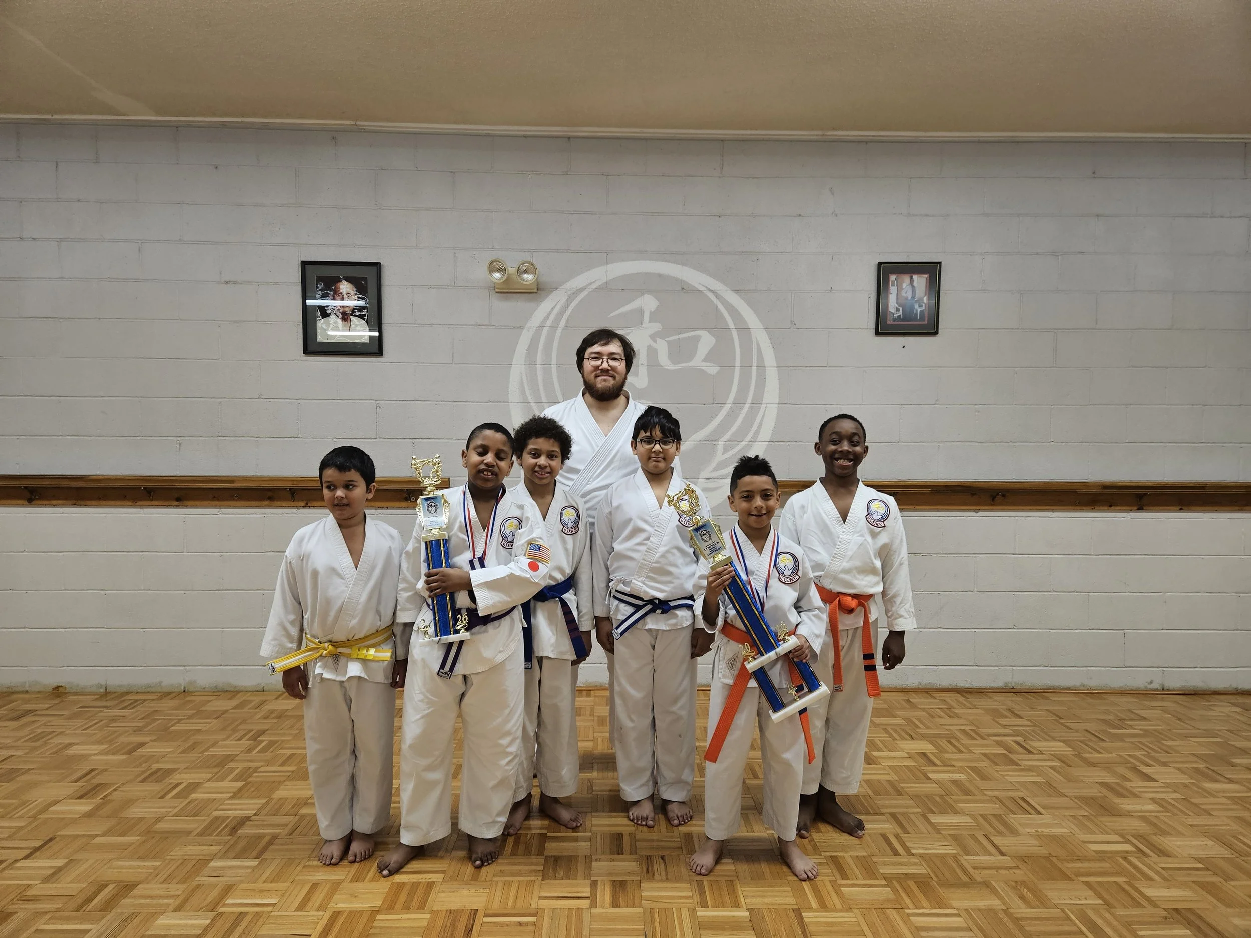Group of young martial artists and their instructor in a dojo, holding trophies and medals after a competition.