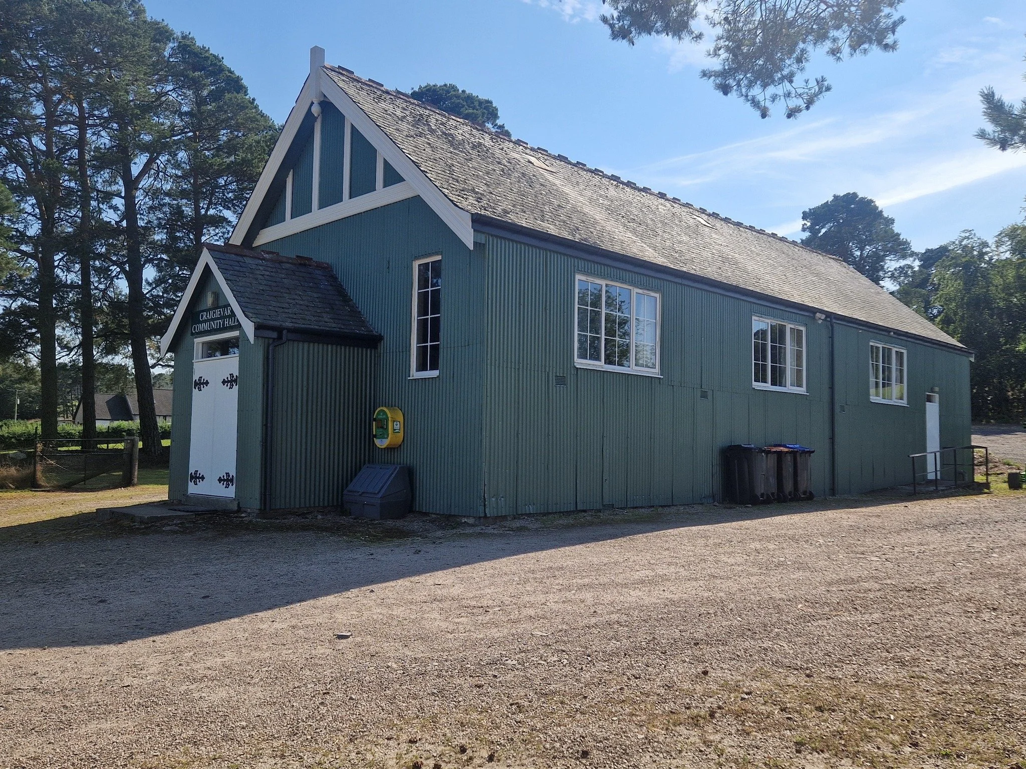 A green wooden community hall with white trim, multiple windows, and a gabled roof, with trash bins outside and a gravel ground in front, surrounded by trees under a blue sky.
