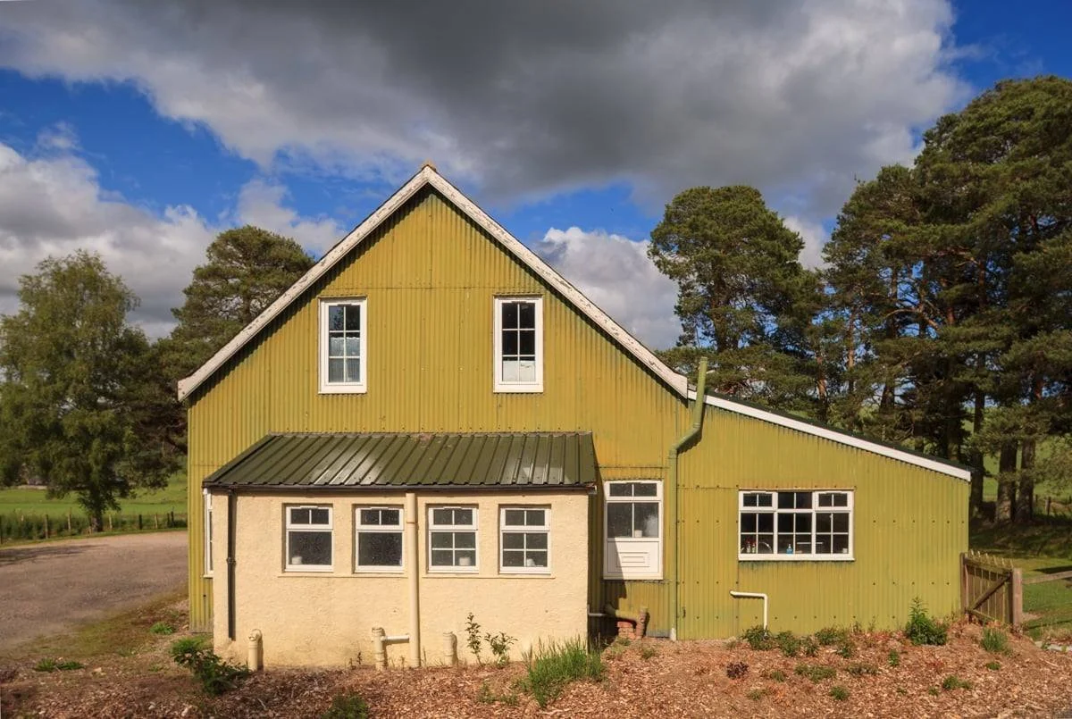 A green village hall with a yellow extension, situated outdoors under a partly cloudy sky, surrounded by green trees and a dirt area in the foreground.