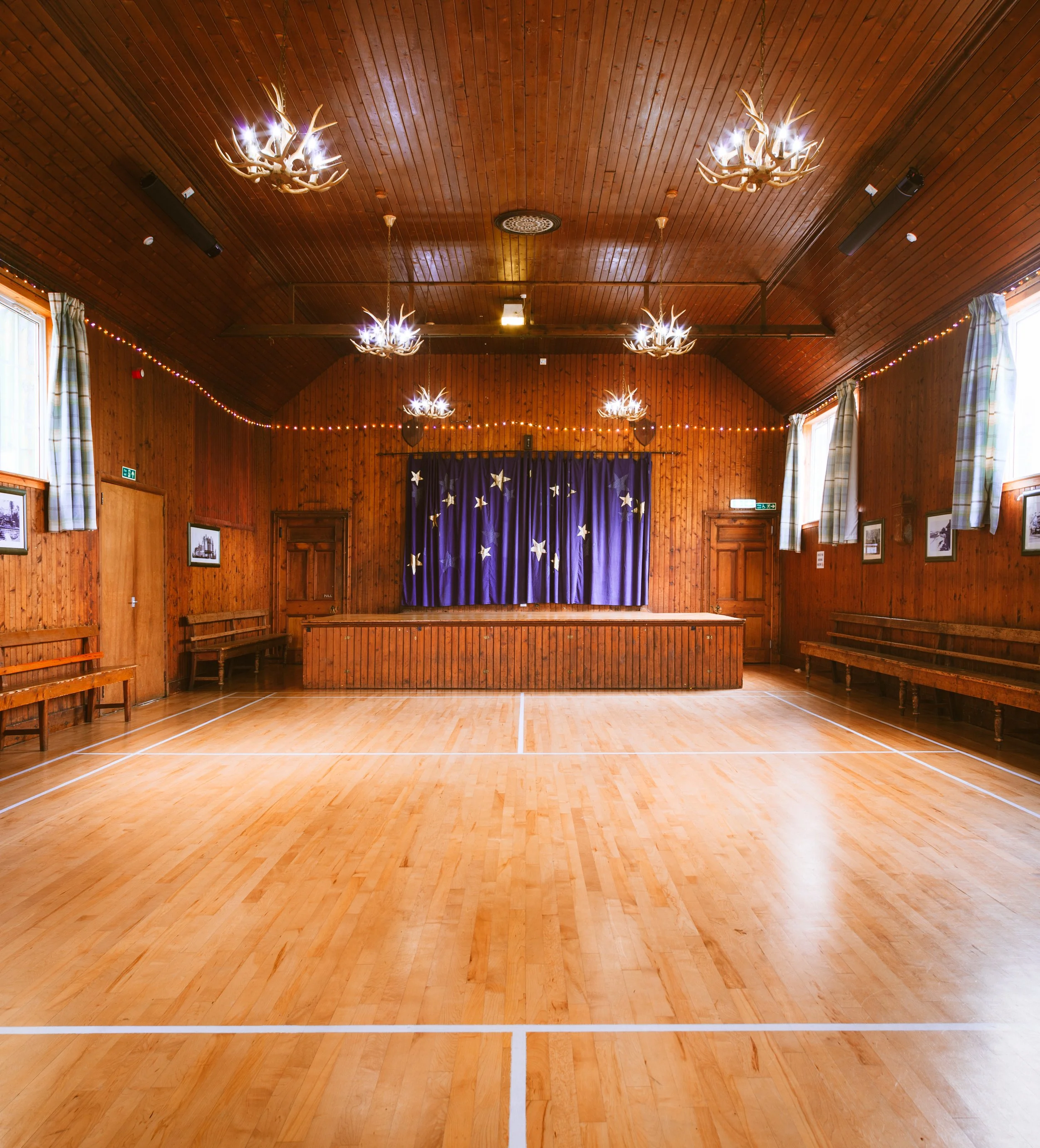An empty wooden hall with a stage at the back, blue starry curtains, and benches along the sides. The ceiling has chandeliers resembling deer antlers, and small string lights are hanging across the room.