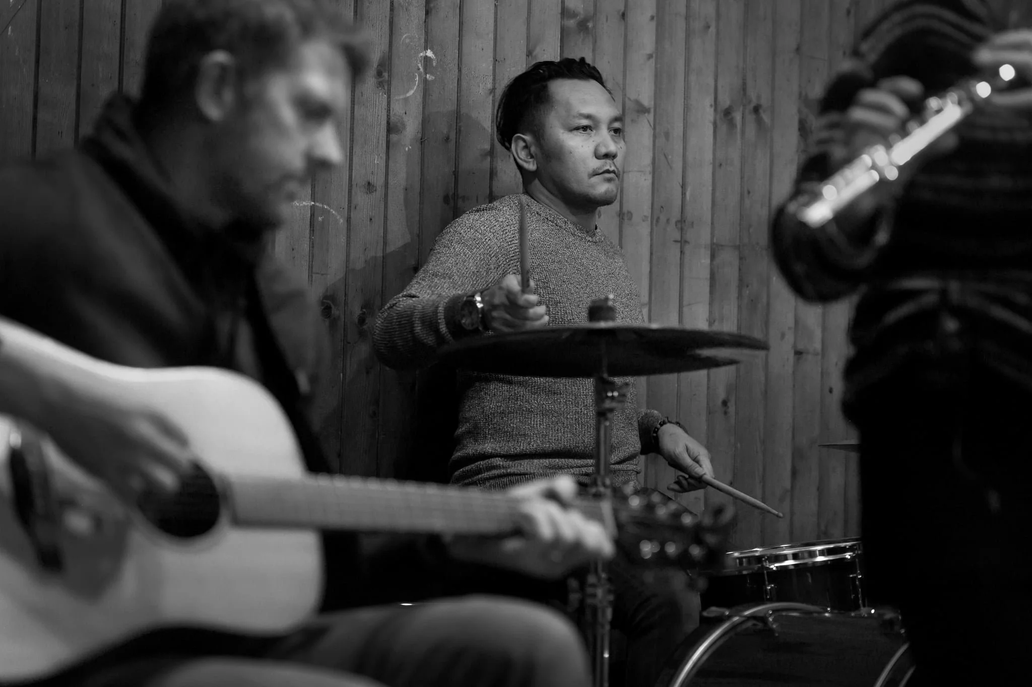 A black and white photo of three musicians, one playing an acoustic guitar, another playing a drum set, and the third with a guitar strap, in a wooden room.