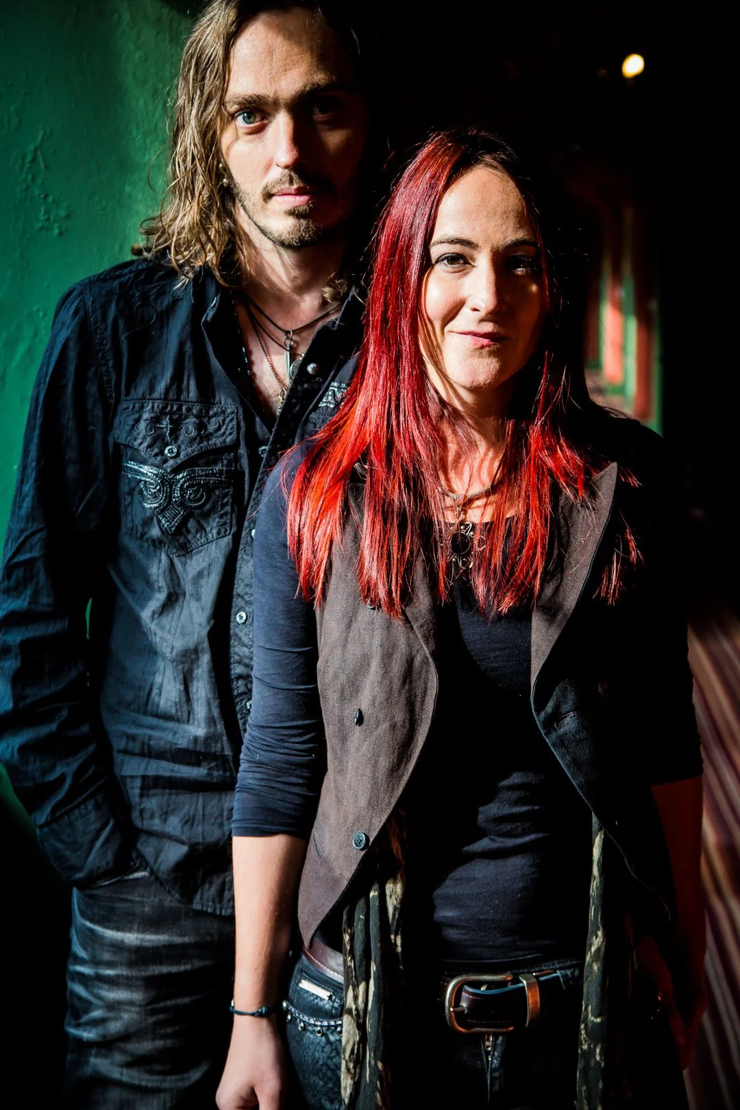 A man with long brown hair stands behind a woman with long red hair, both dressed in dark clothing, posing together in a dimly lit, atmospheric setting.