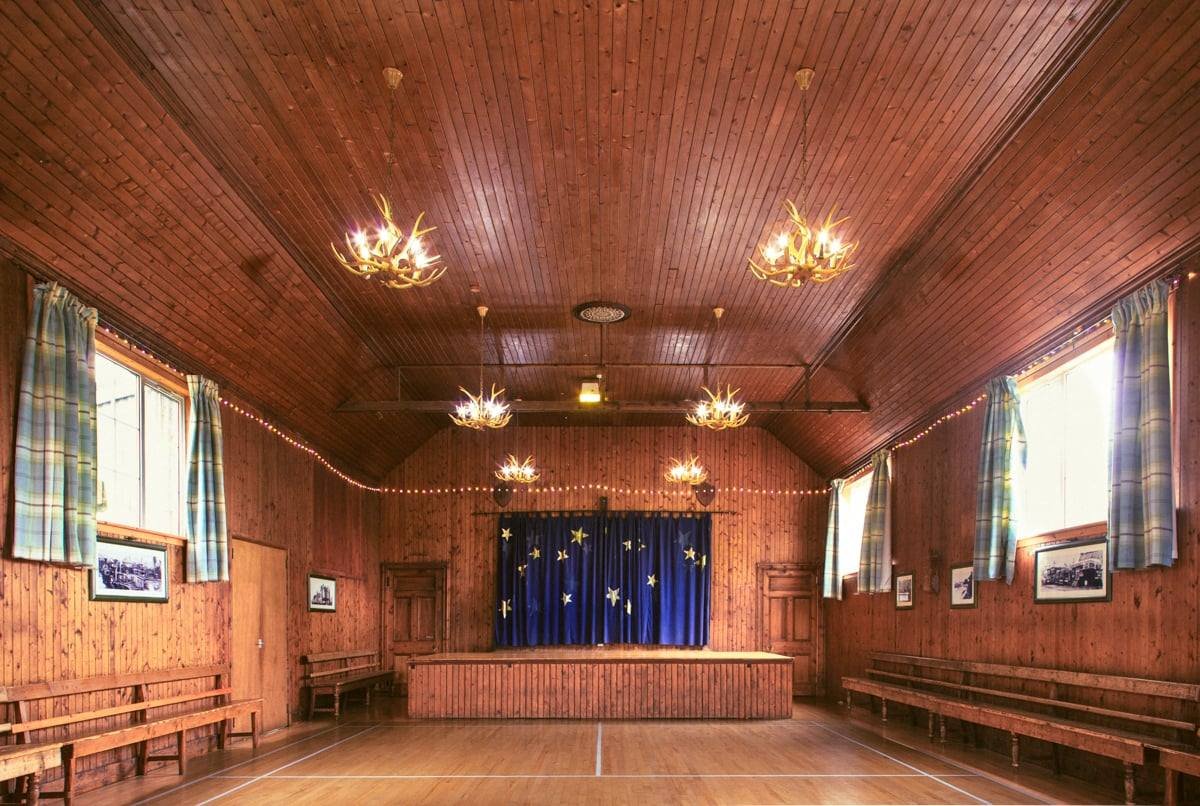 Interior of a wooden hall with a small stage and blue starry curtain, striped curtains on windows, and wooden benches along the walls.