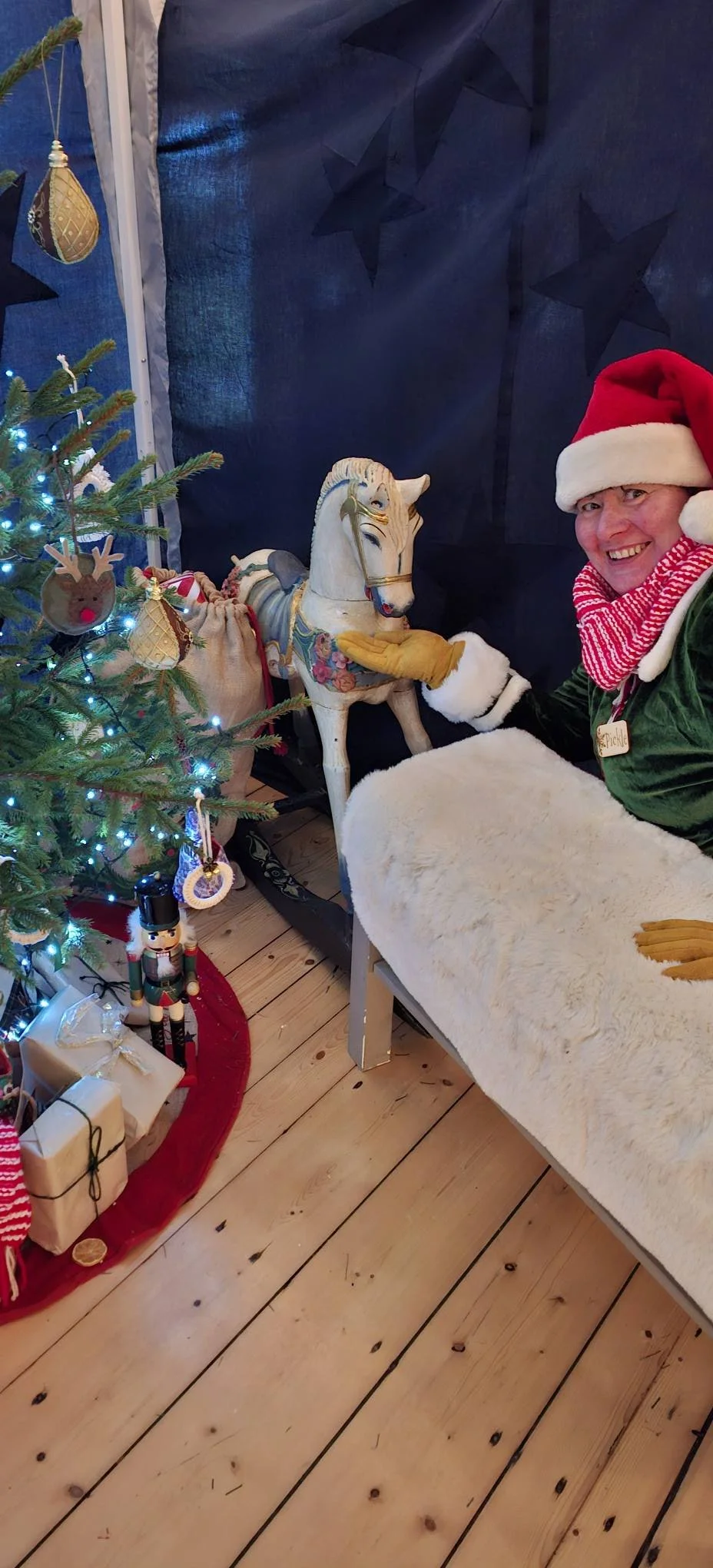 A person dressed as Santa Claus smiling and posing with a vintage-style rocking horse next to a decorated Christmas tree with lights and ornaments, in a cozy, festive setting.