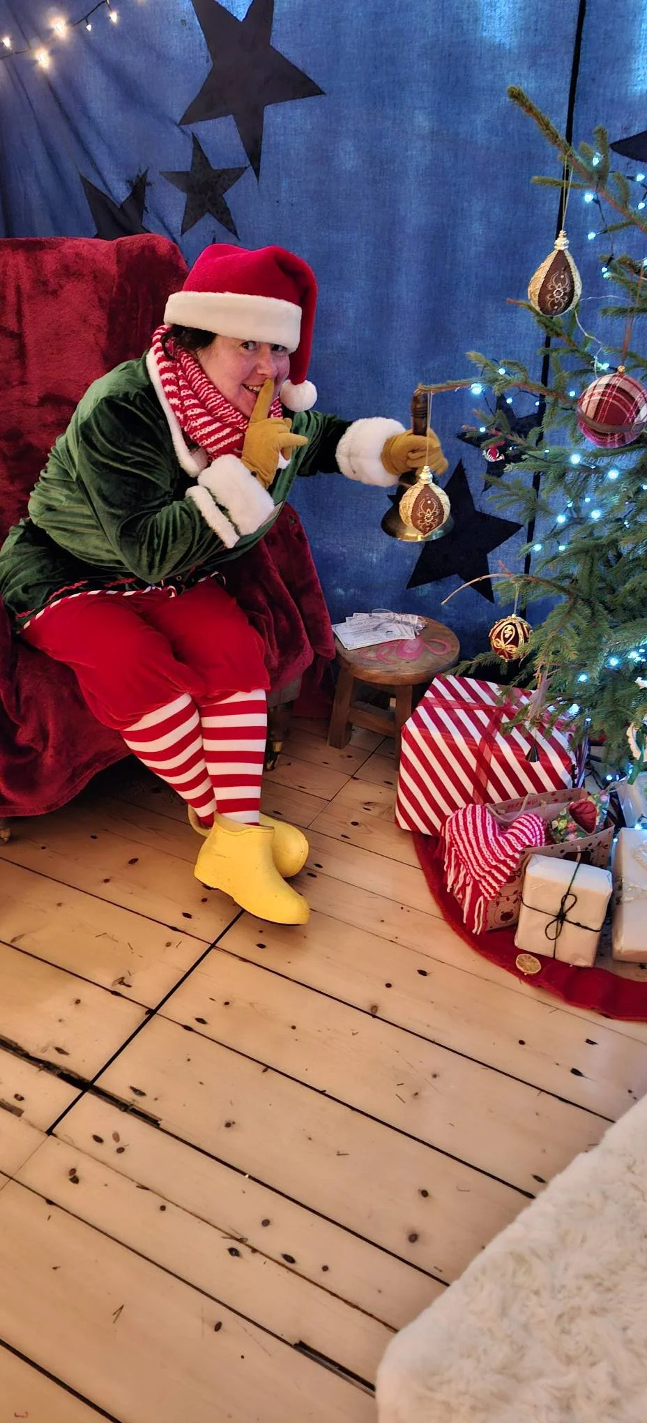 A person dressed as an elf sitting on a red armchair with a Santa hat, yellow gloves, red and white striped tights, and yellow shoes, holding a bell and making a shushing gesture. There is a decorated Christmas tree with presents underneath, and blue