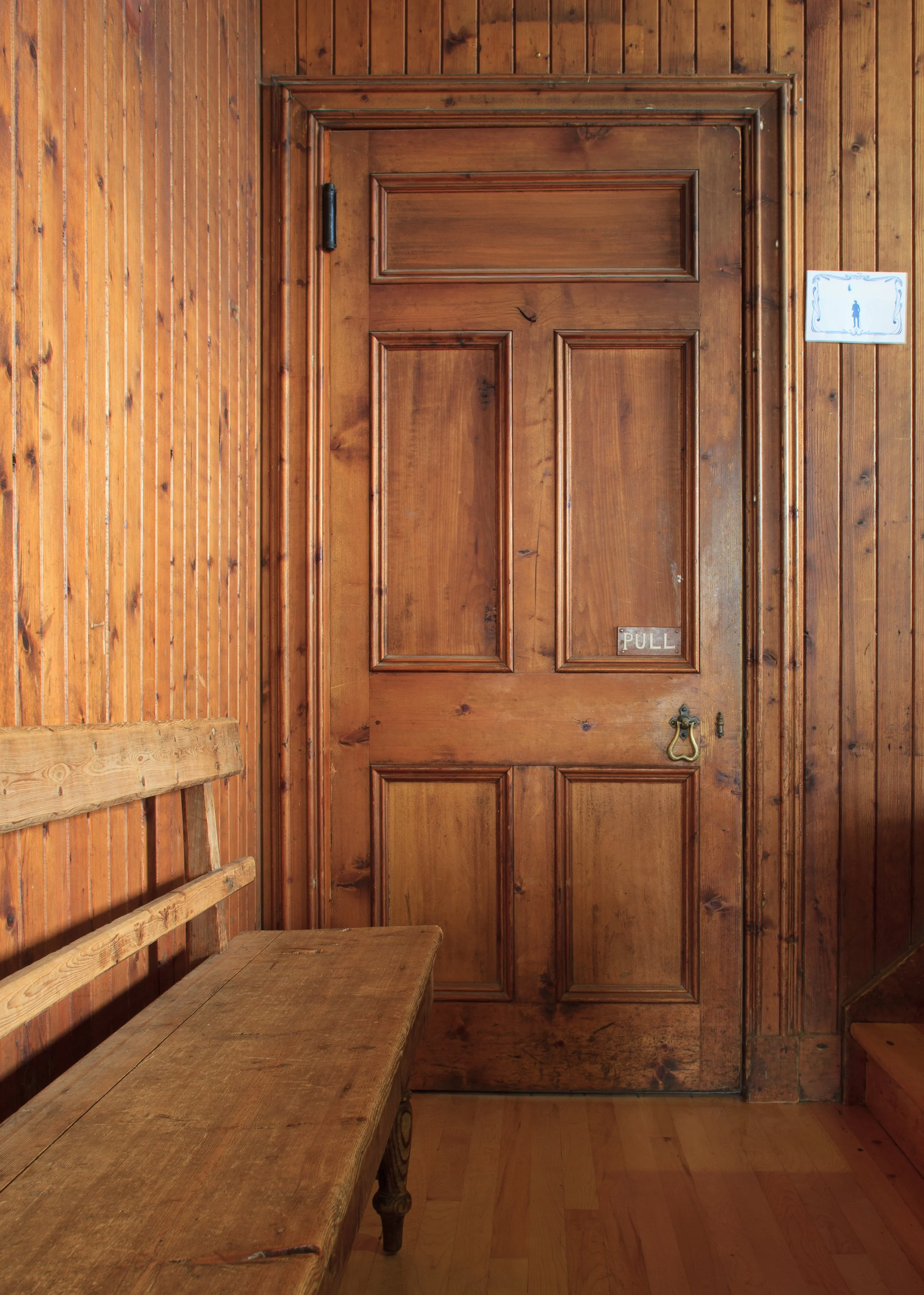 A wooden door with a brass handle and a small sign that says 'PULL'. The door is set in a wood-paneled wall, with a wooden bench to the left.