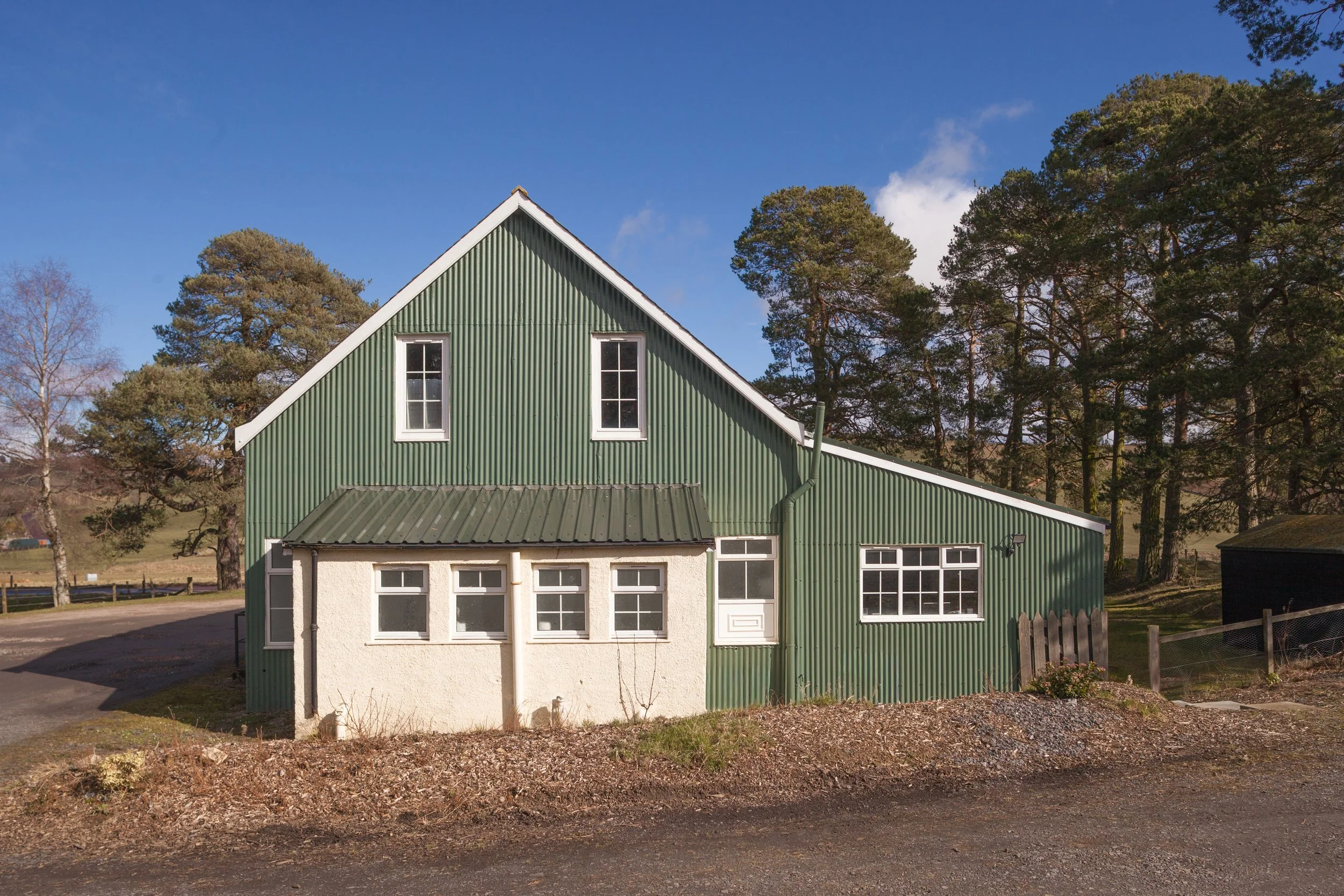 A green, corrugated metal house with a sloped roof, several white-framed windows, and a small white door, set against a background of trees and a blue sky.