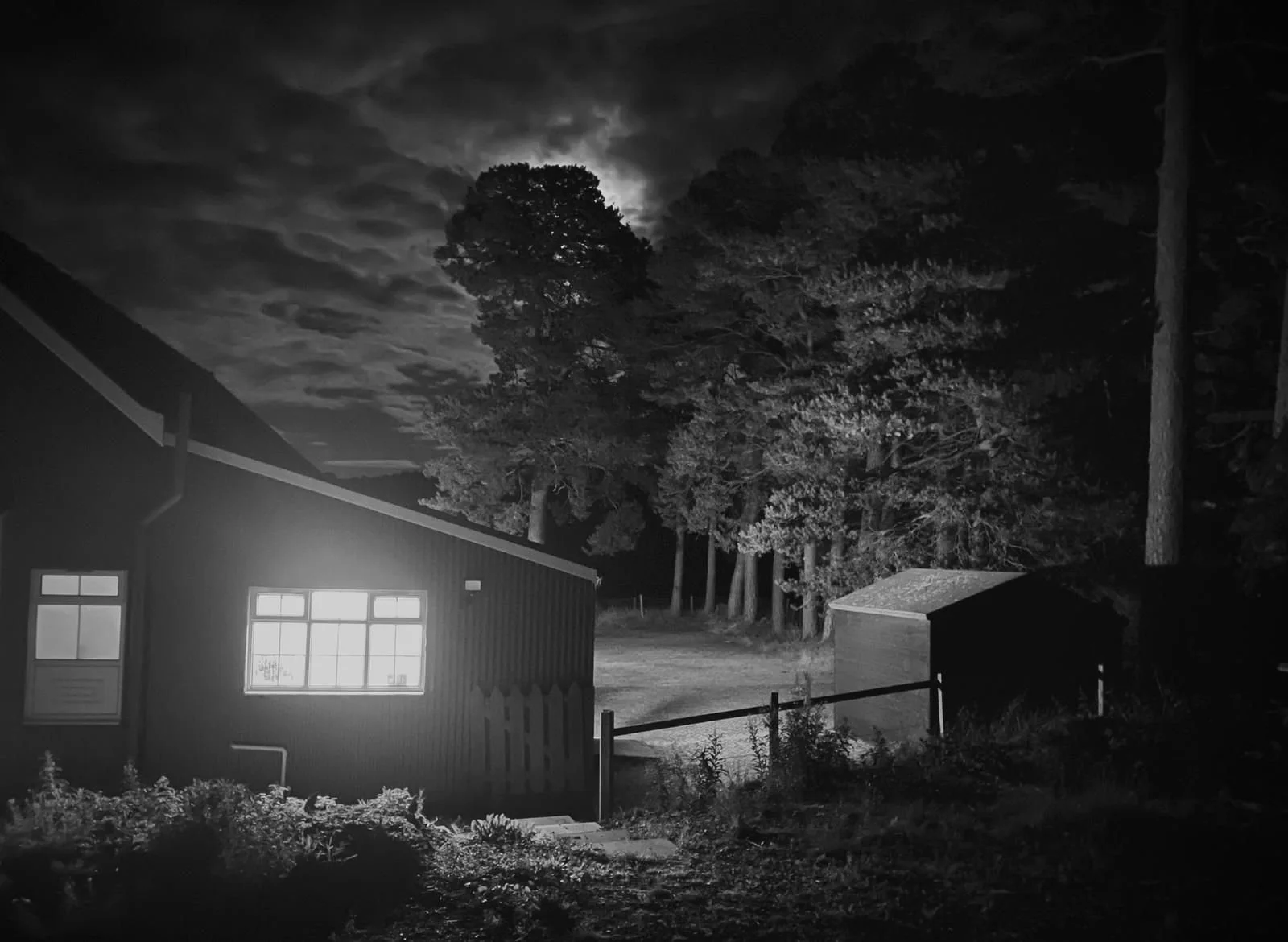 Night scene in black and white with a house illuminated from the inside, trees in the background, and a cloudy sky. This is slightly spooky