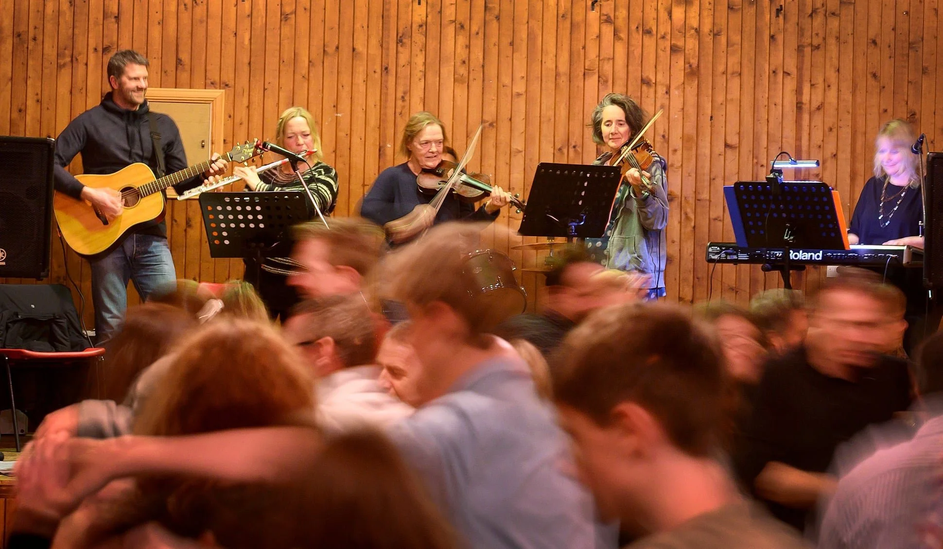 A band of five musicians performing on stage with a wooden wall background, including a guitarist, two violinists, a keyboardist, and a woman singing or playing the violin. Audience members are dancing or hugging in front of the stage.