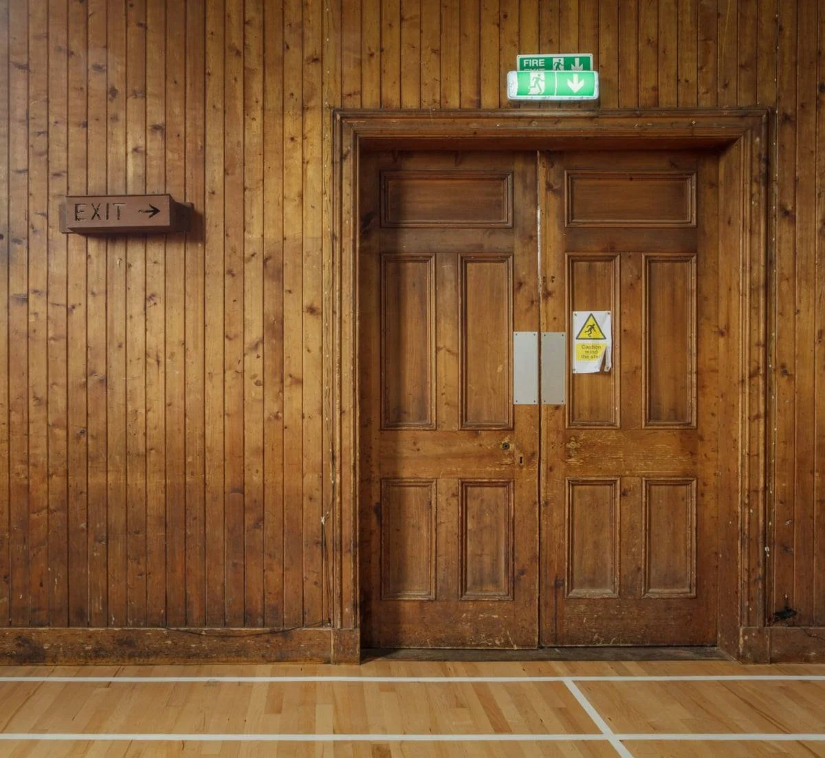 Wooden wall with a large wooden door, exit sign, and fire exit signs above. A caution sign is on the door.