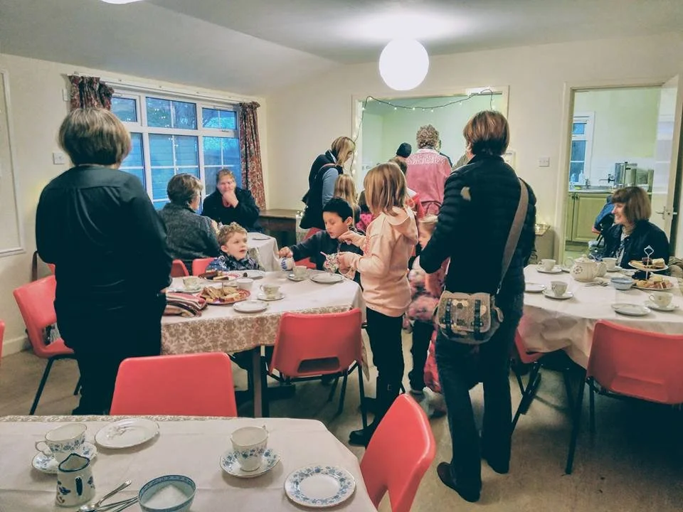 Family and friends gathered around dining tables with tea cups, plates of food, and decorations in a cozy room for a social or celebratory meal.