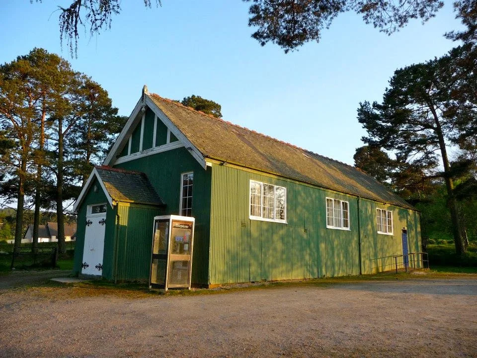 A green barn with a sloped roof, white trim, and a telephone booth outside, set in a rural area with trees and a gravel driveway.