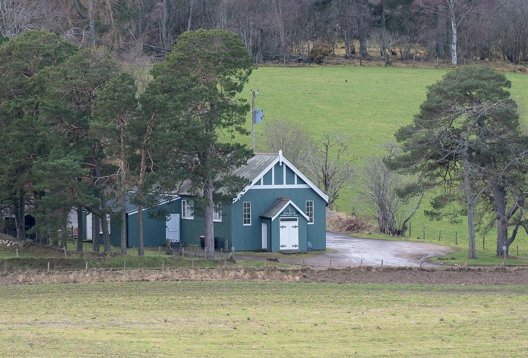 A rural scene with a blue community hall building surrounded by trees, grass, and hills.
