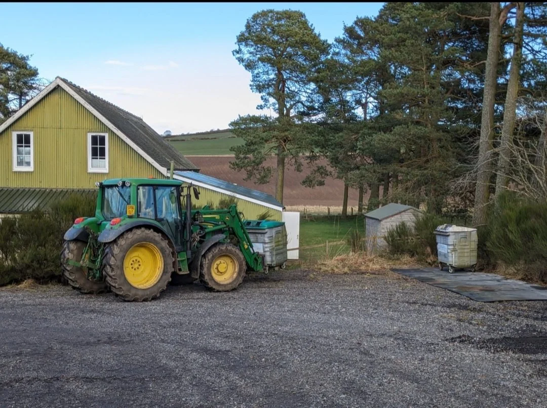 A green tractor parked on a gravel surface near a yellow house with a sloped roof, surrounded by trees and open fields in the background.
