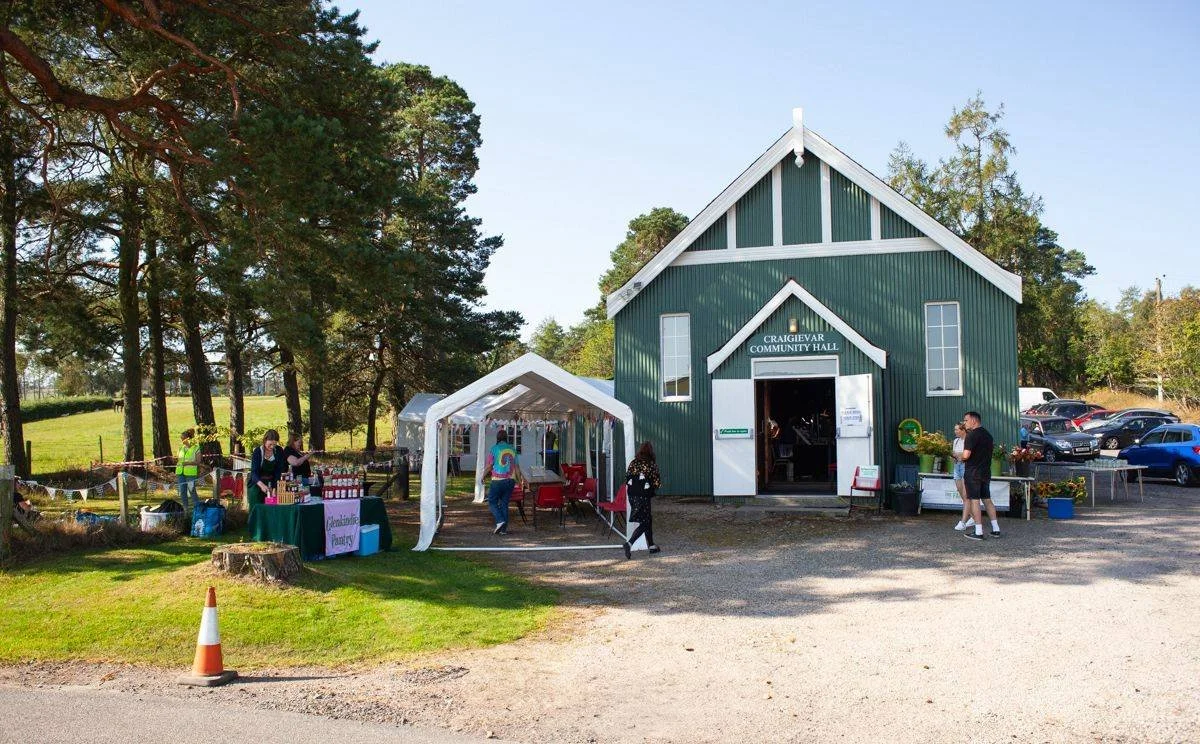 A community event outside a green wooden community hall with a white sign that reads "CRAIGDIEVAR COMMUNITY HALL." There is a tent set up with people browsing items on tables, and several people walking around or standing nearby. Cars are parked to t