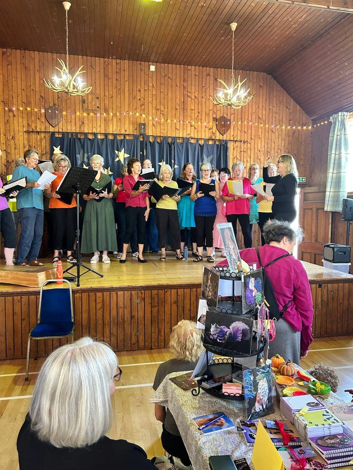 A group of women singing in a choir on a stage in a wooden hall during a community event, with some seated at tables selling artwork and crafts.