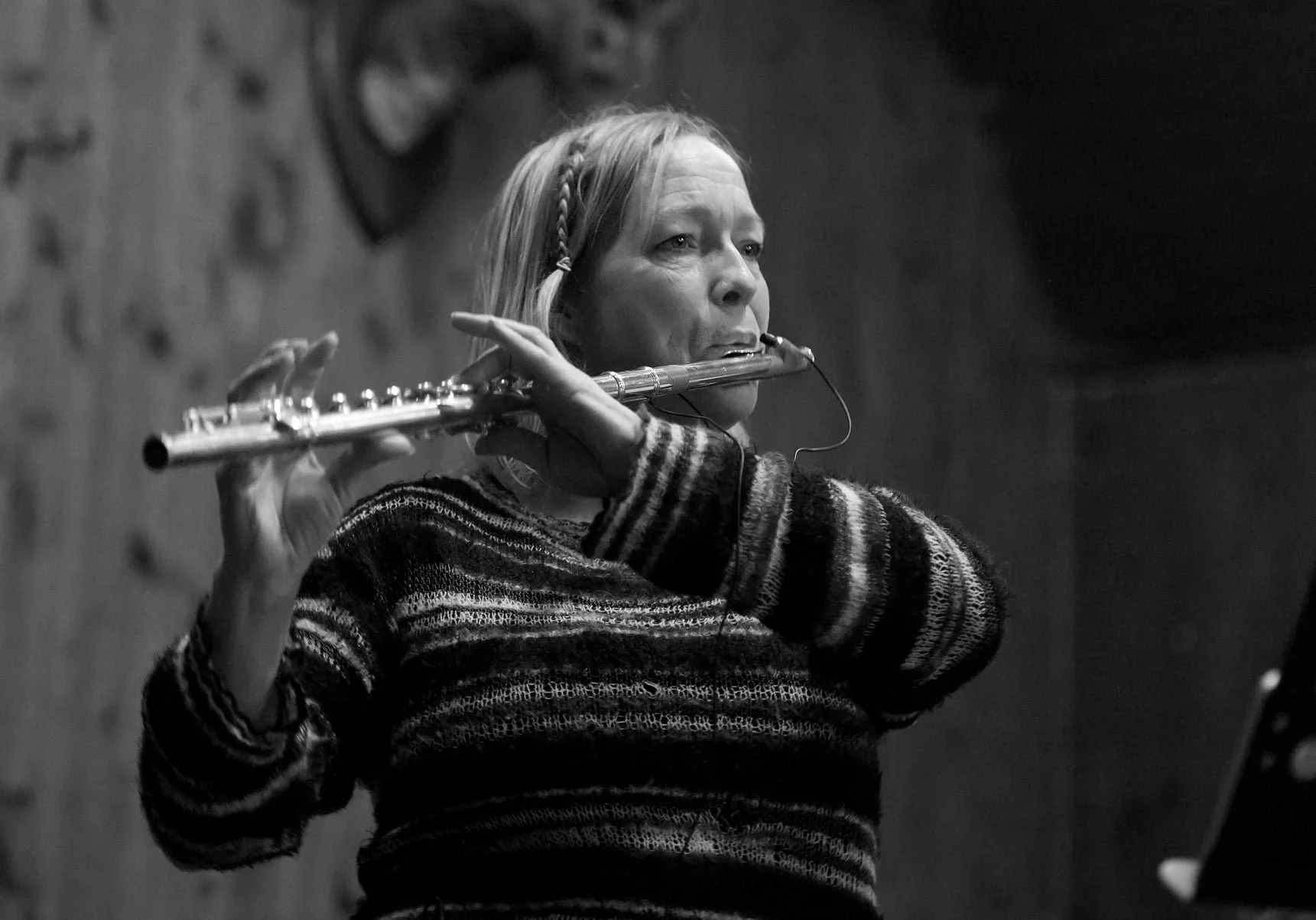 Black and white photo of a woman playing a flute, wearing a striped sweater and a braided headband, in a studio or practice room with wooden walls.
