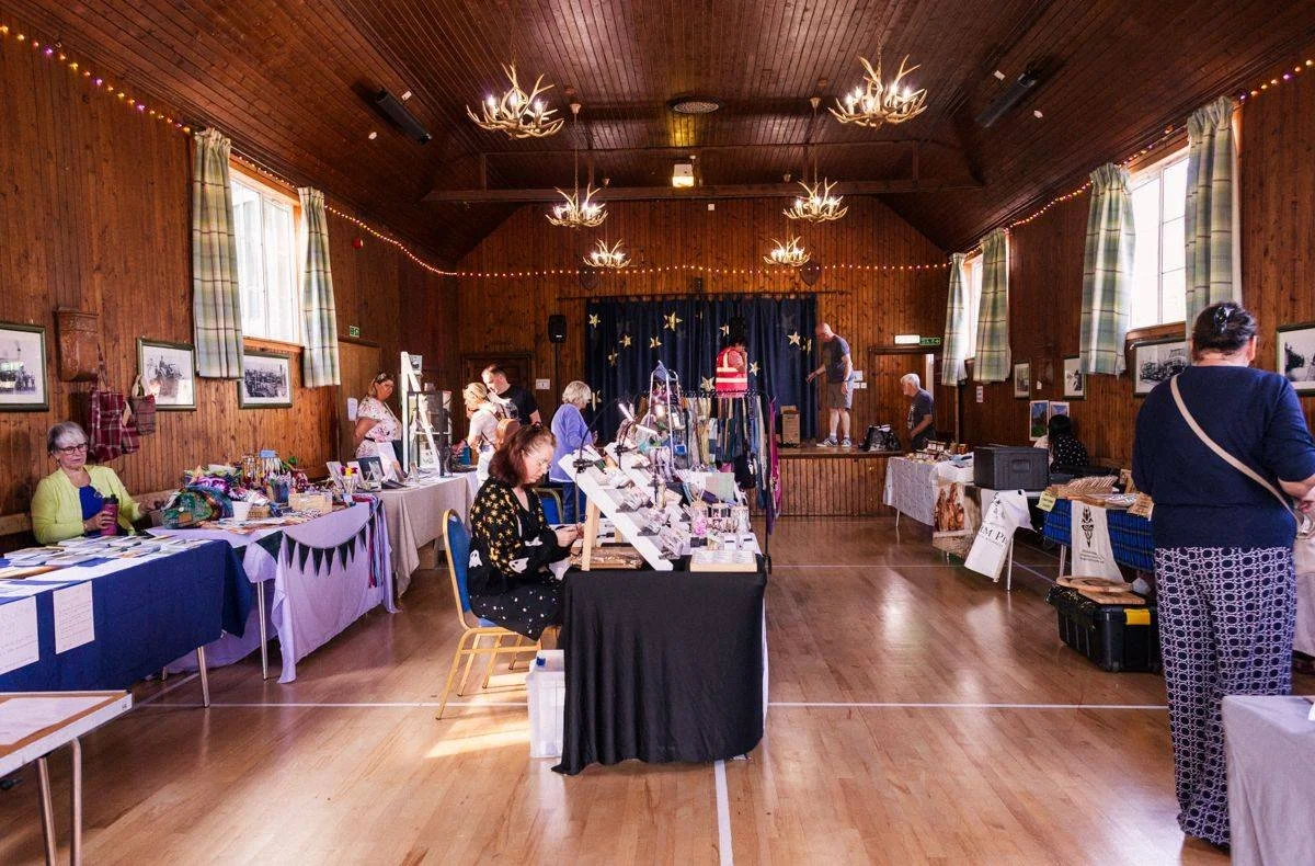 A craft fair inside a wooden hall with vendor tables displaying handmade items and jewelry. Several people browse and chat, with a small stage at the back and festive lighting.