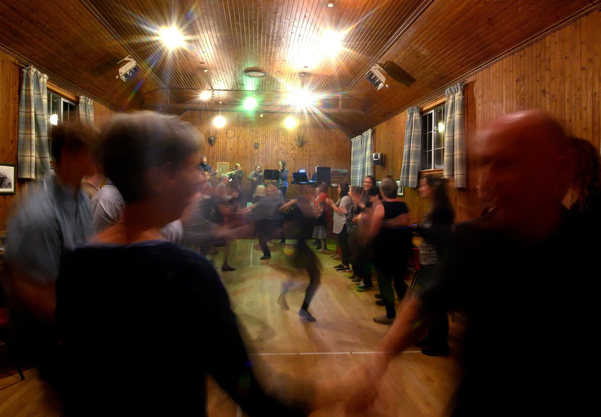 People dancing in a wooden-floored room with wood-paneled walls, windows with plaid curtains, and mounted animal heads, while a band plays on stage in the background.