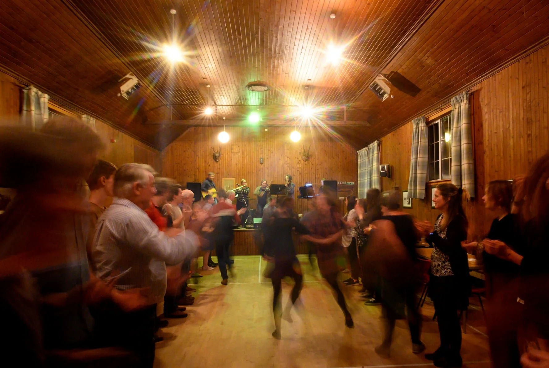 People dancing and socializing in a wooden-floored hall with a live band on stage, under bright ceiling lights.