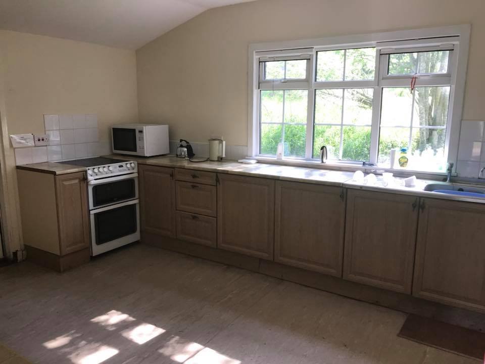 A small kitchen with wooden cabinets, a white stove, a microwave, a window above the sink, and some dishes and cleaning supplies on the countertop.