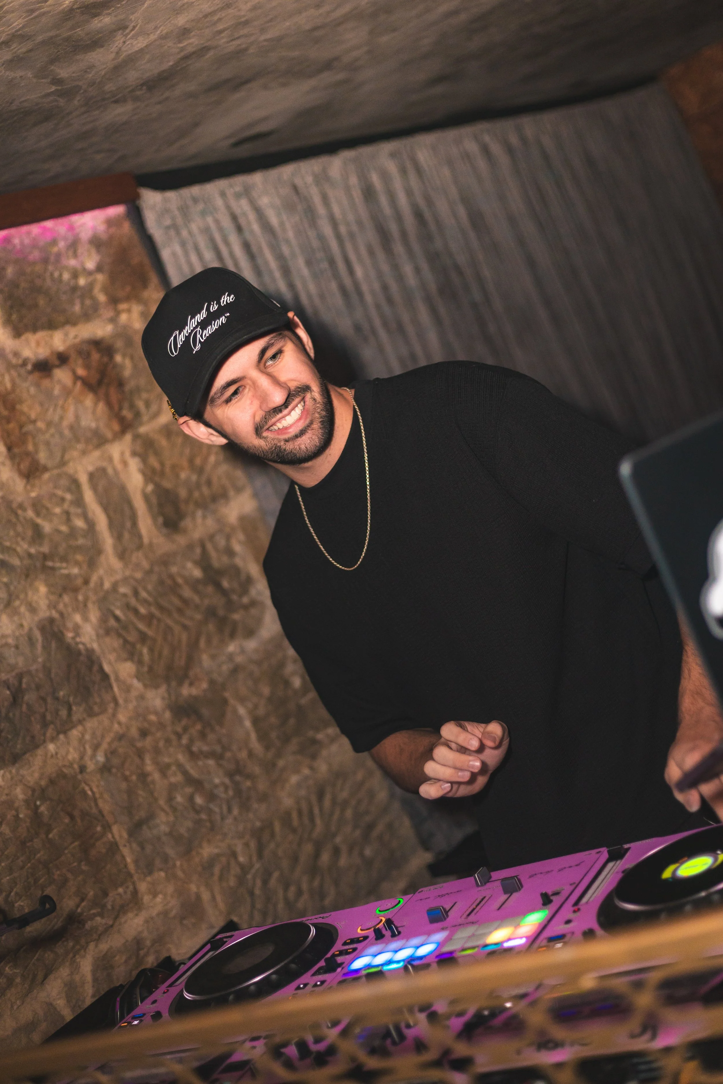 A man smiling and looking to the side, wearing a black shirt, a chain necklace, and a black cap that says 'Auckland is the Reason,' standing next to a DJ turntable setup.