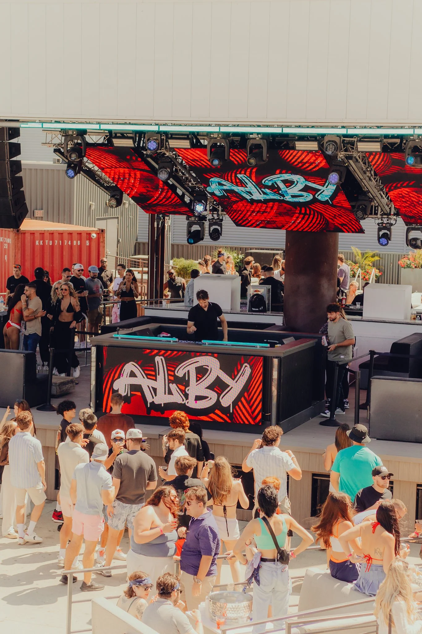 Crowd of people at an outdoor party with Alby performing EDM and house music on a stage with a large screen and colorful lights.