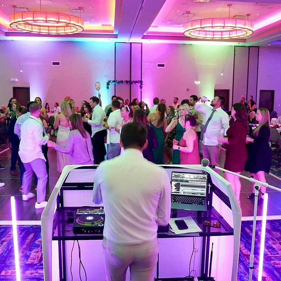 A DJ is playing music at a party or wedding reception, with guests dancing in a brightly lit banquet hall decorated with colorful lights and chandeliers.