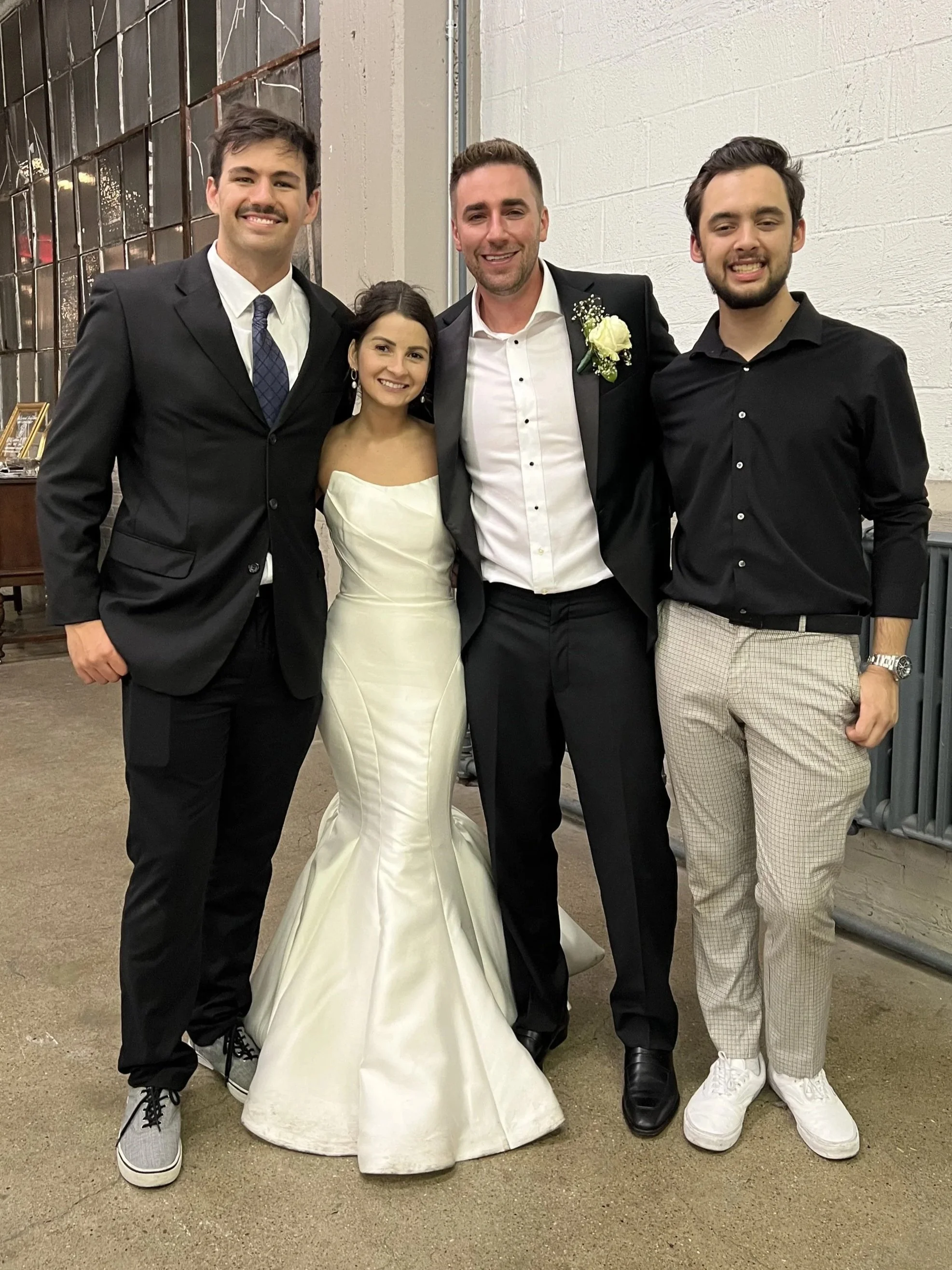 Four young adults dressed in formal attire, standing together and smiling at an indoor event, with a rustic industrial background.