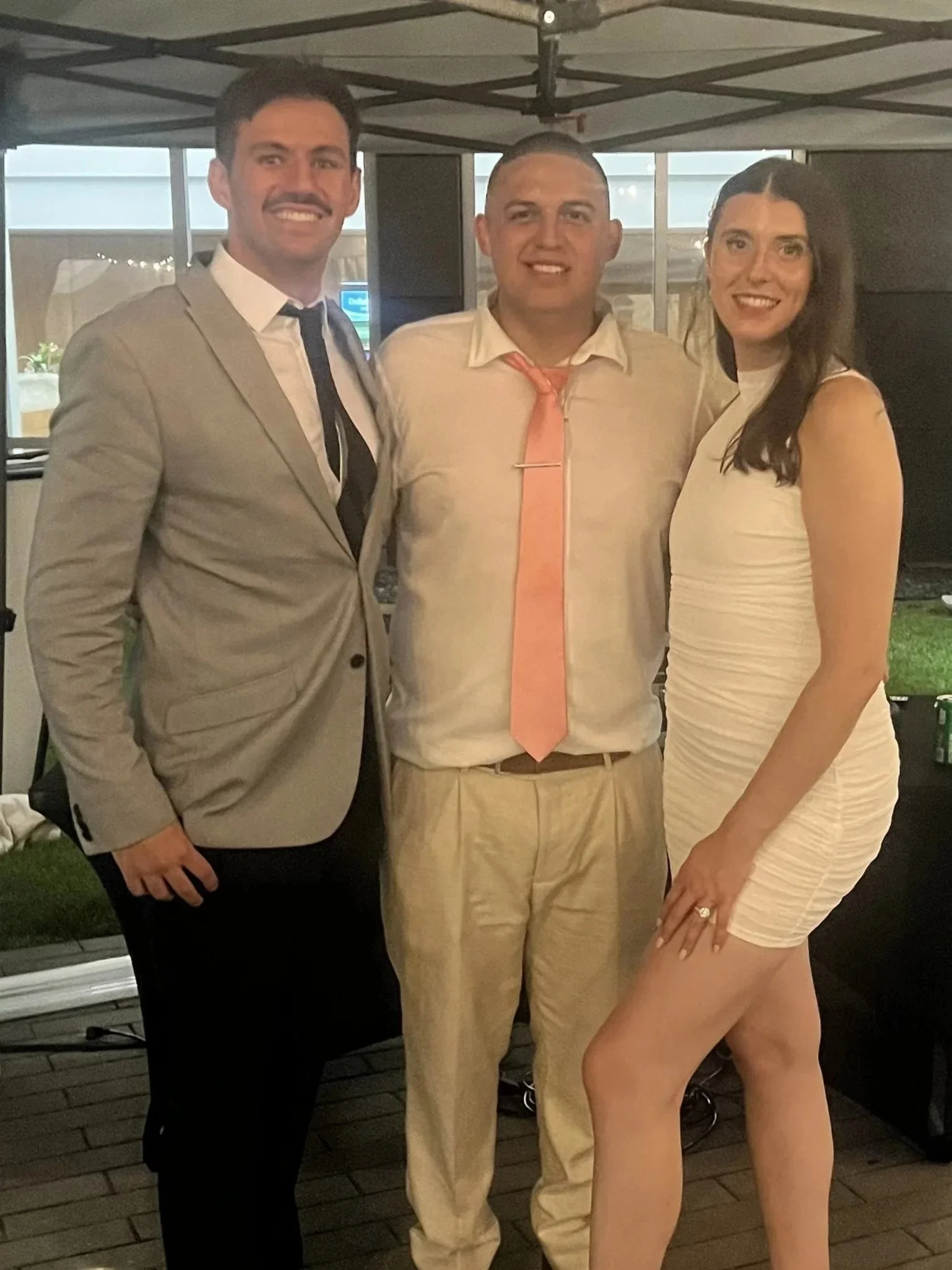 Three people standing together outdoors at night, under a canopy, smiling at the camera. The man on the left wears a light gray suit and black tie, the man in the middle wears a white shirt and tie, and the woman on the right wears a tight, sleeveles