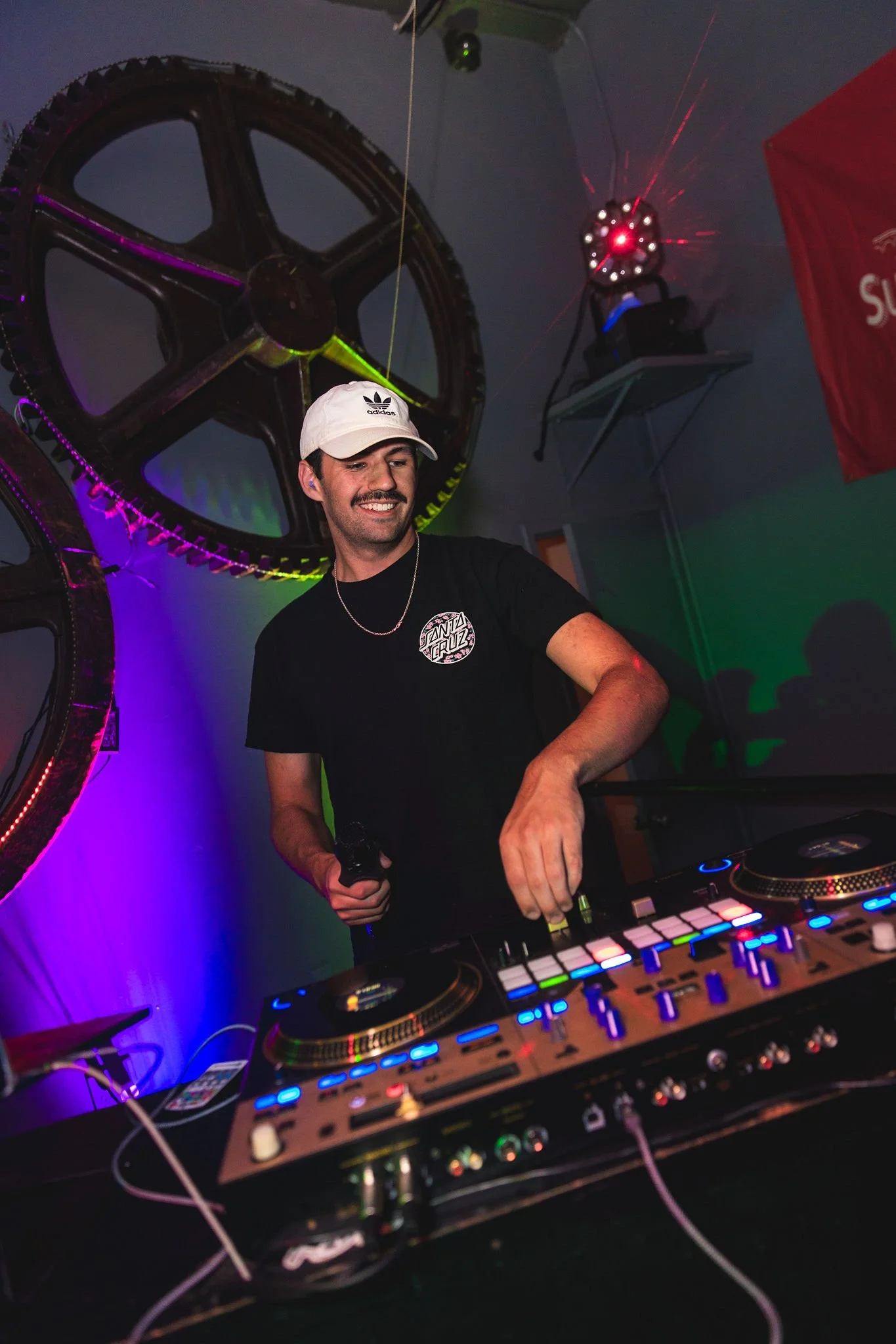 A DJ with a white cap, black T-shirt, and necklace is mixing music at a club with colorful lighting and large bicycle gears as decoration on the wall behind him.