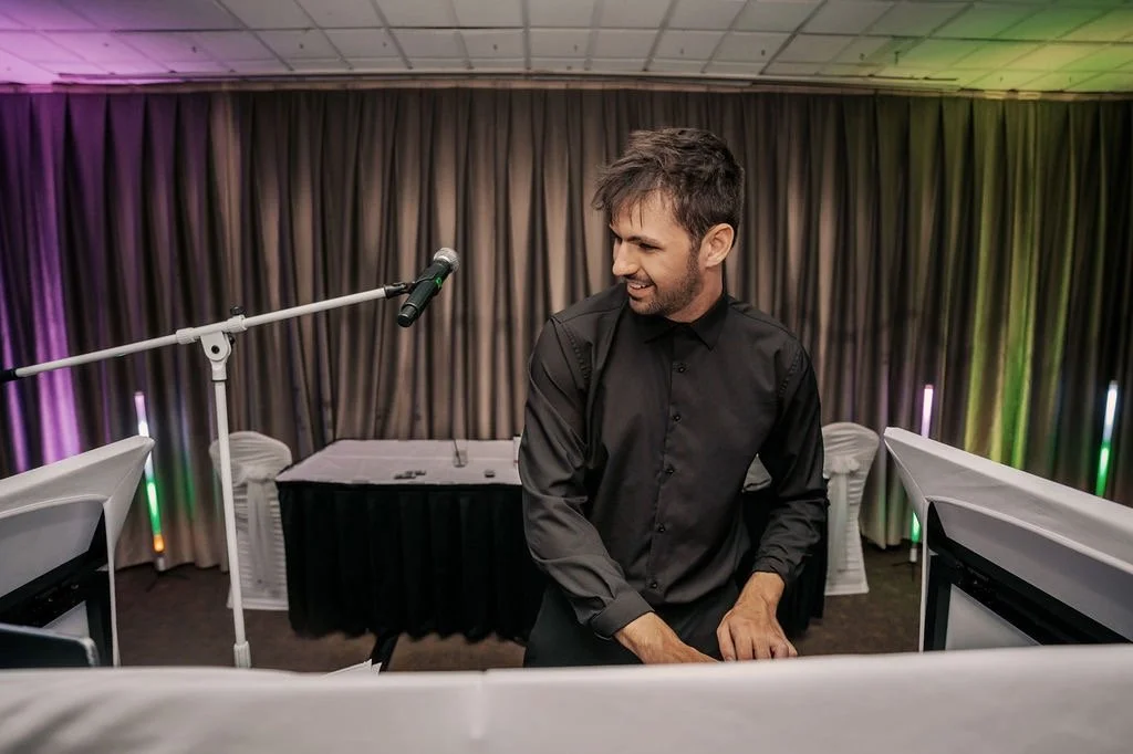 A man in a black shirt is standing behind a DJ booth, smiling, with a microphone stand in front of him, in a room with curtains and colorful lights.