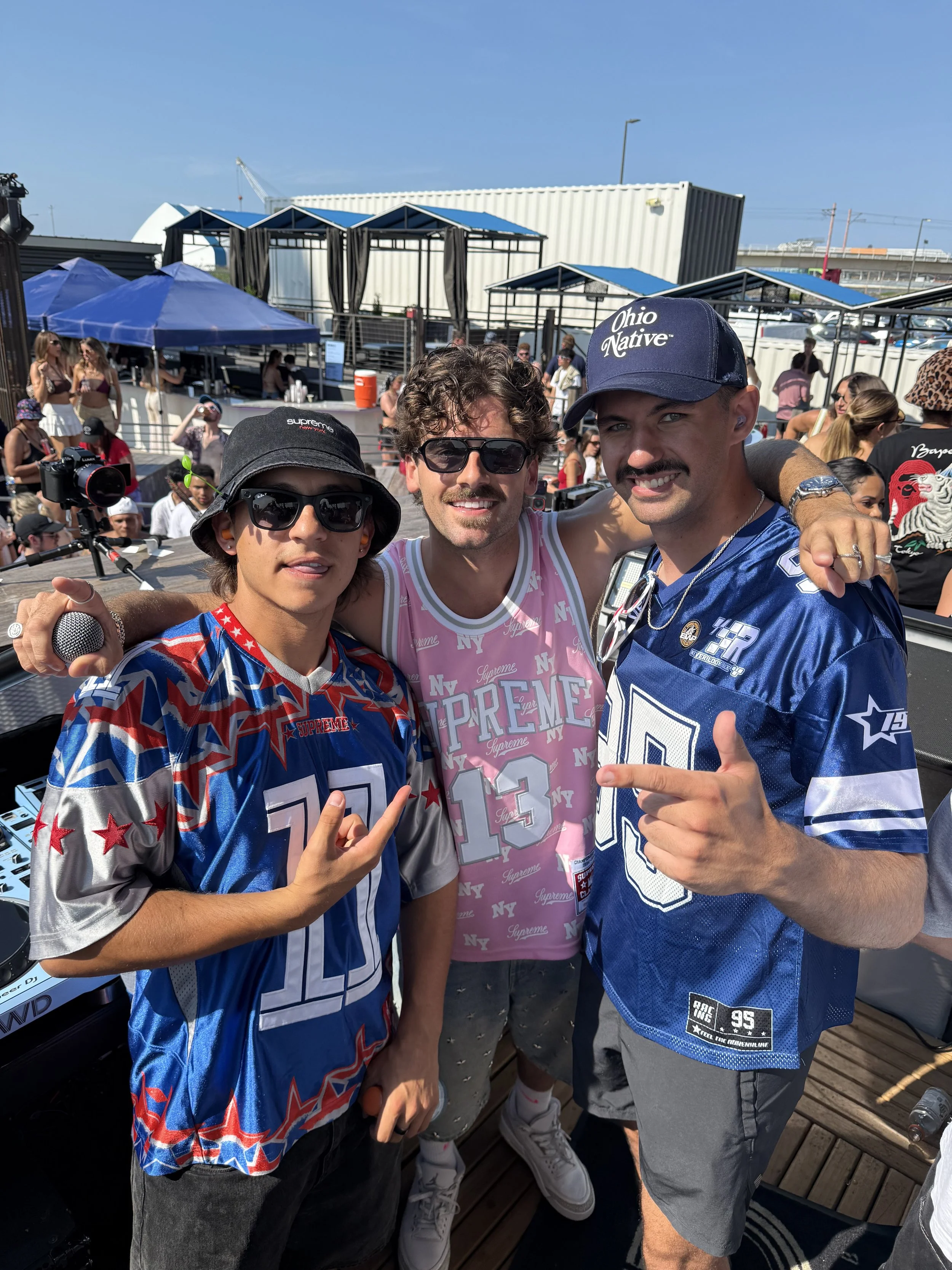 Three men posing together at an outdoor event with a crowd and tents in the background, smiling and making hand gestures.