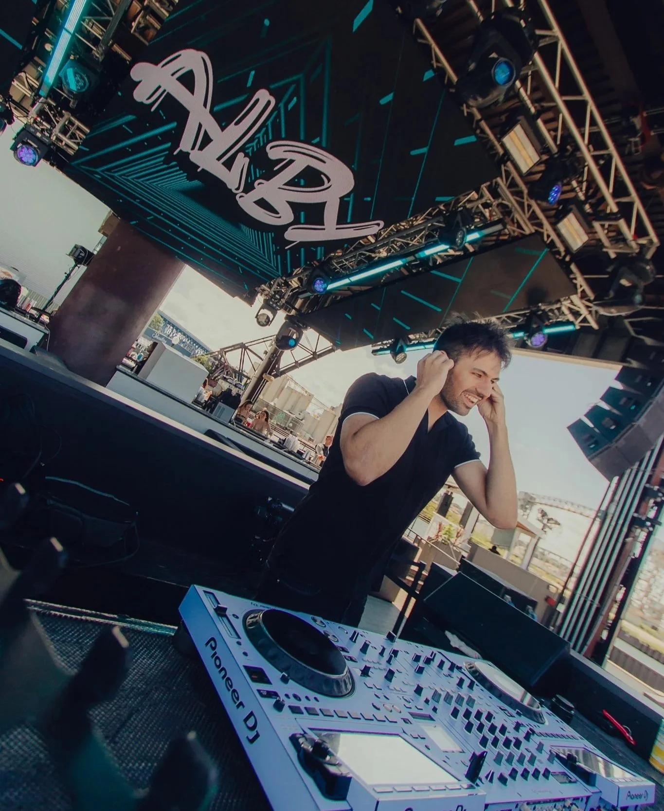 Alby with headphones smiling on an outdoor stage, with DJ equipment labeled Pioneer DJ, under a canopy with a large illuminated sign.