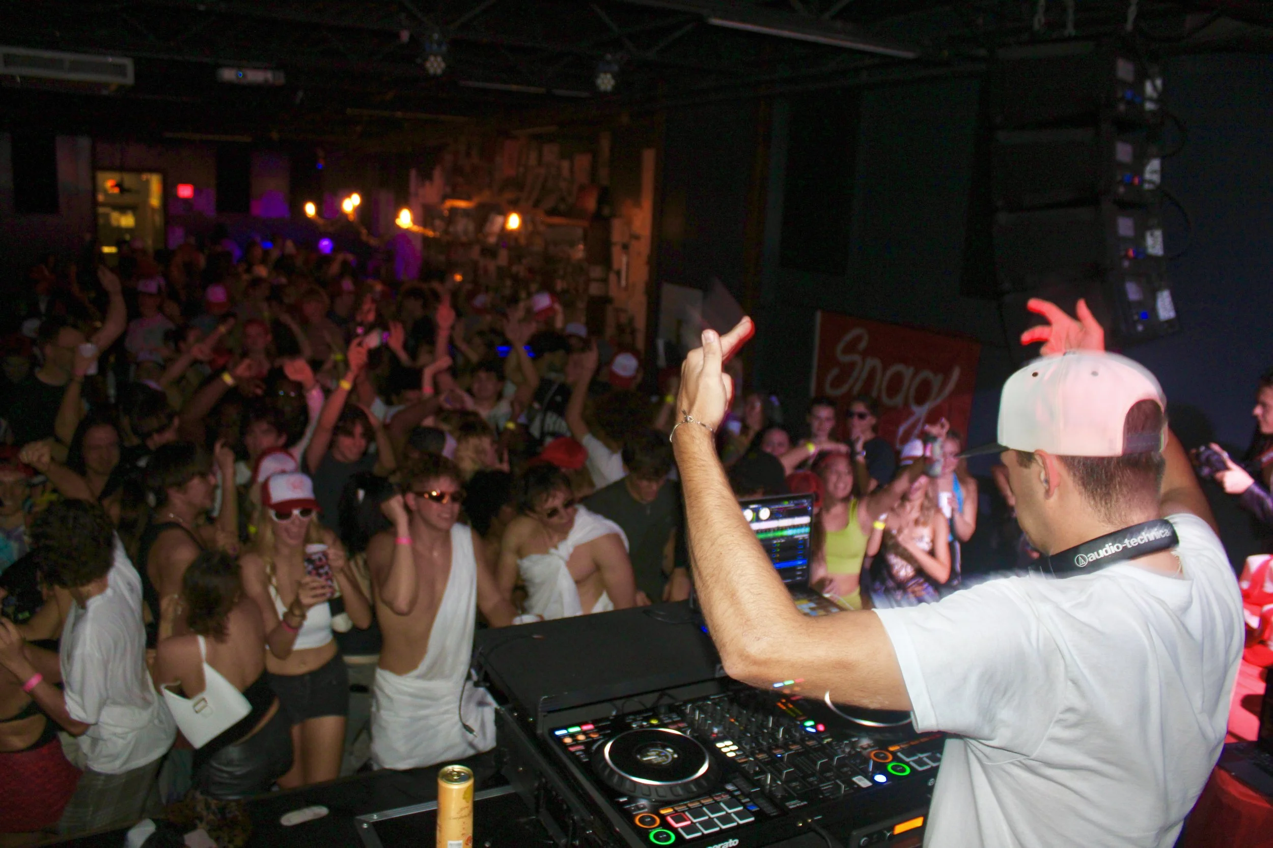 DJ performing at a crowded indoor music event, wearing a white cap backwards, with a laptop and DJ equipment, and a lively audience dancing and enjoying the music.