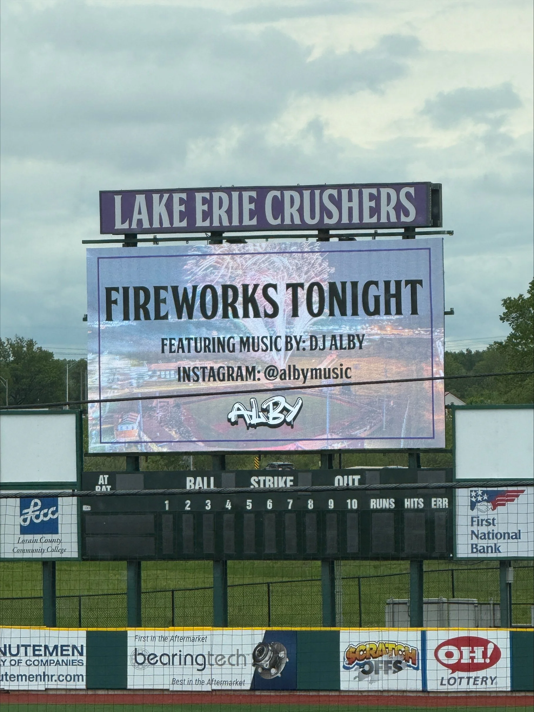 Billboard at a baseball field advertising fireworks event with music by DJ Alby, at Lake Erie Crushers. The scoreboard shows, "Fireworks Tonight," with social media handle @albymusic, and logos for First National Bank and other sponsors.