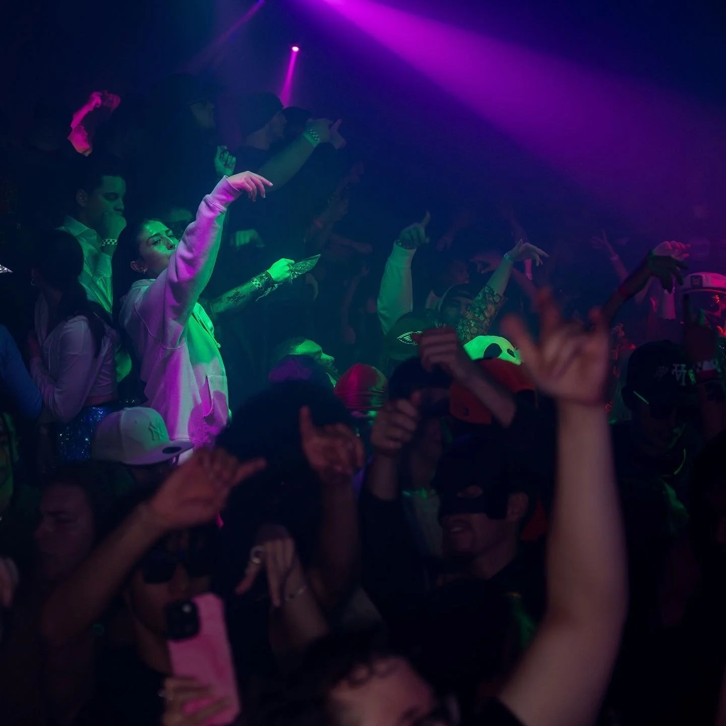 Crowd of people at a concert or nightclub with neon purple and green lighting, some raising their hands and taking photos.