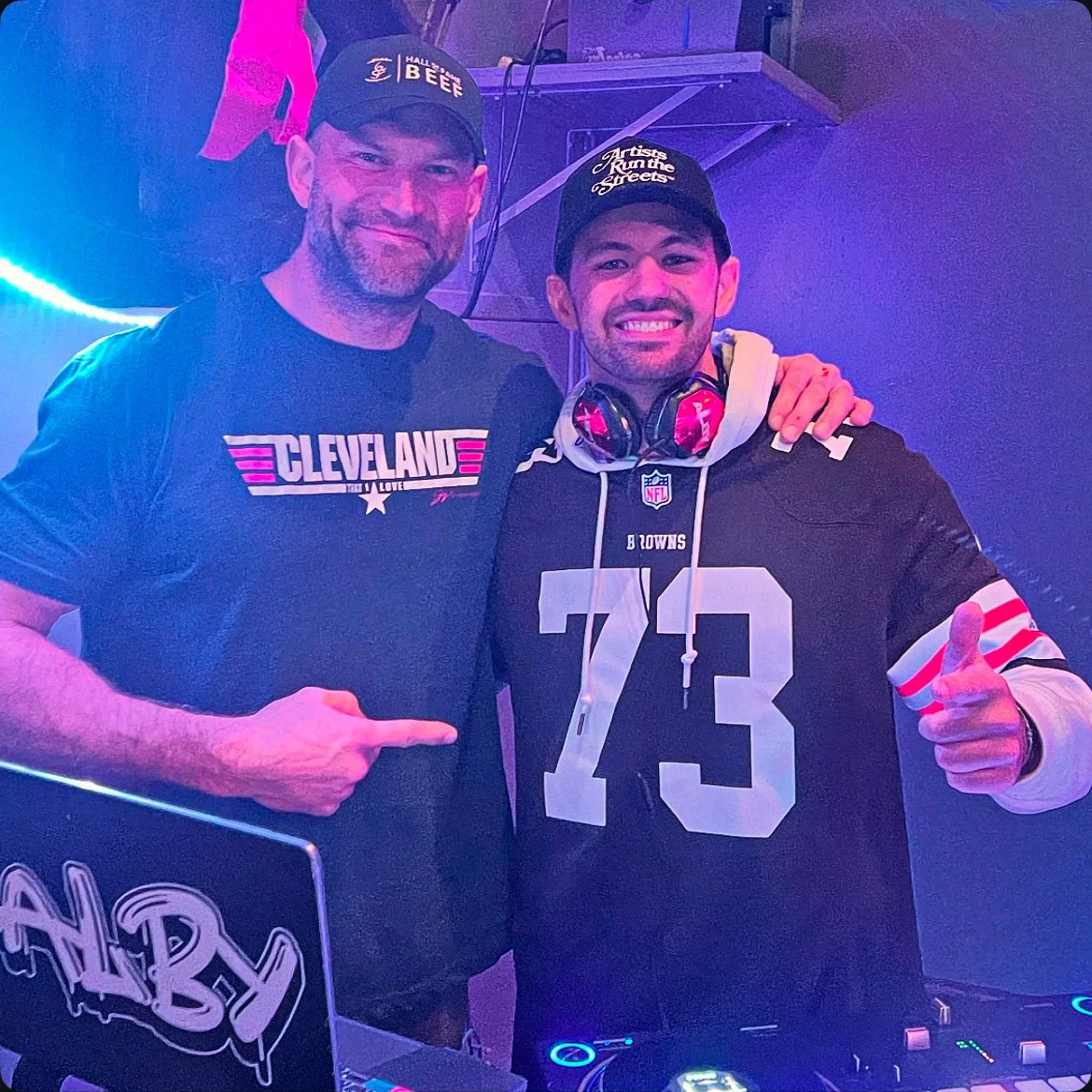 Alby with former Browns player Joe Thomas. Joe Thomas is wearing a Cleveland shirt and a cap, while Alby is wearing a black NFL jersey with the number 73, a cap, and headphones around his neck.