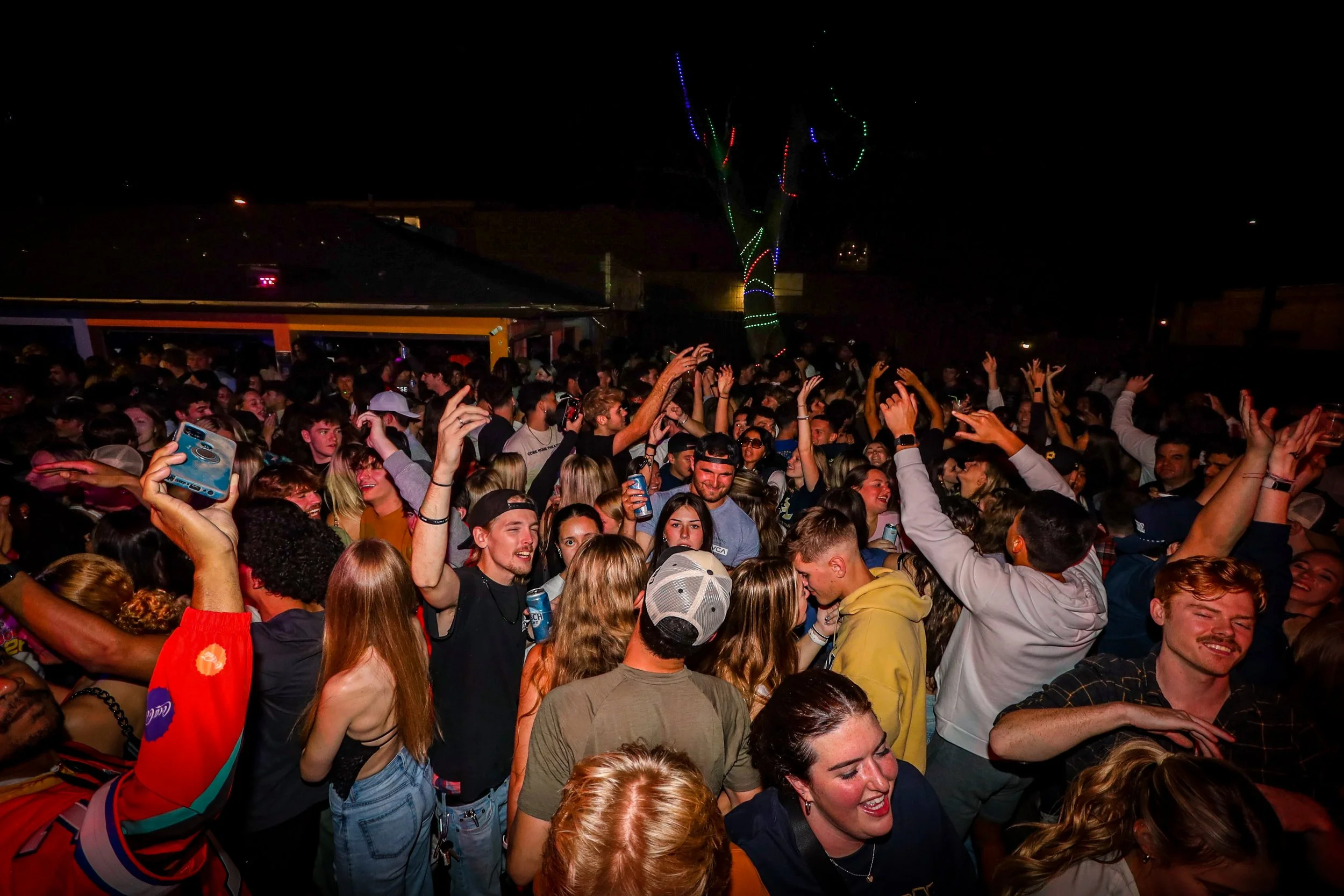 Crowd of young people dancing and enjoying a night outdoor party with a lit tree in the background.