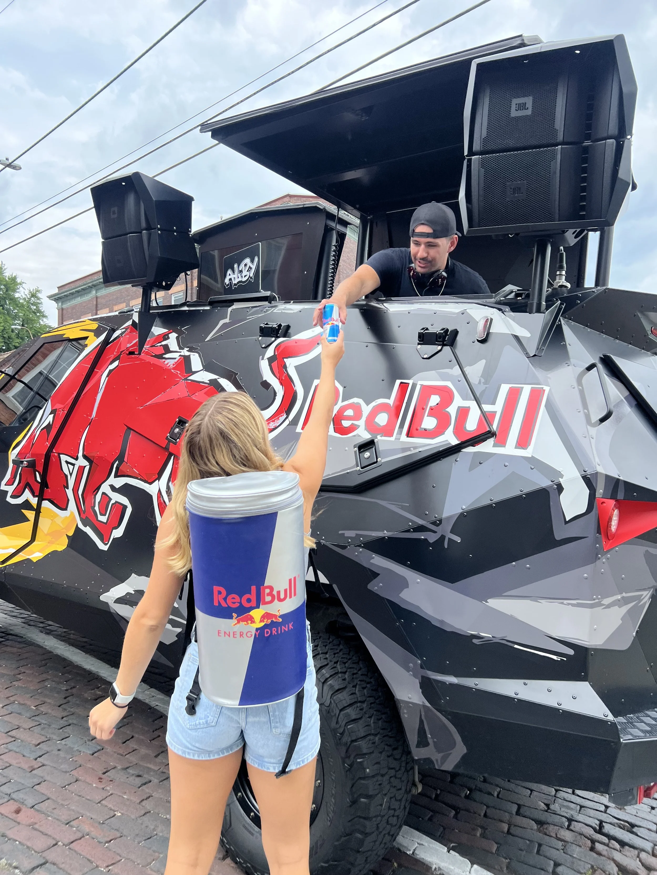 A woman dressed in a Red Bull energy drink costume handing a can to Alby in a Red Bull armored vehicle.